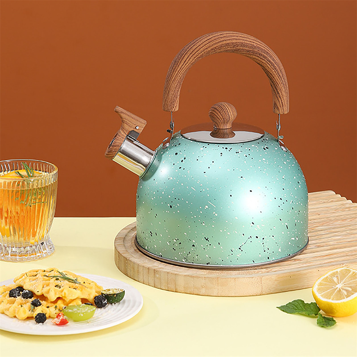 Ein teal-farbener Wasserkocher mit Holzgriff und Deckel. Auf einem Tisch sind auch Lebensmittel zu sehen.