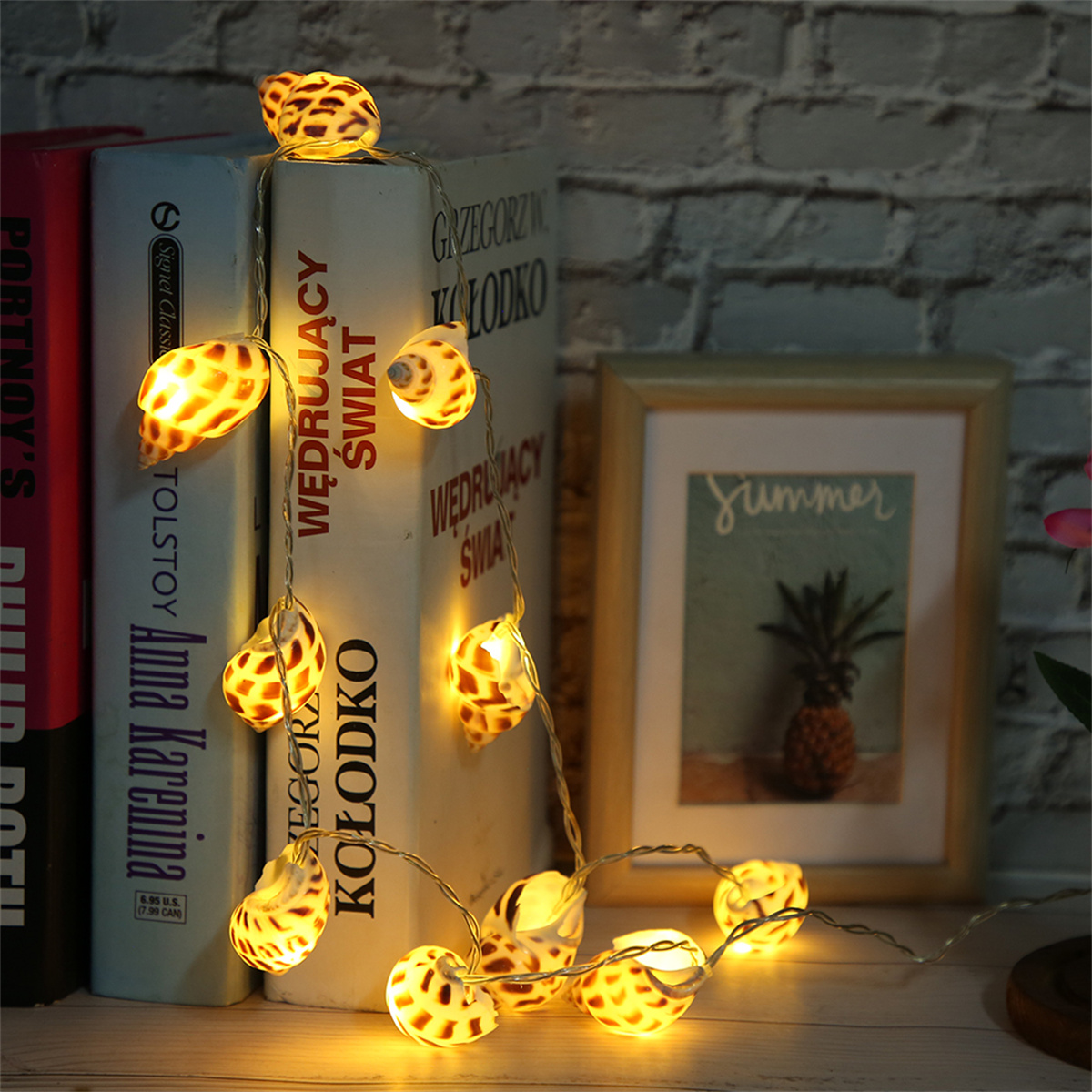Eine Kette aus Muschellichtern neben Büchern und einem Bilderrahmen mit einer Ananas.