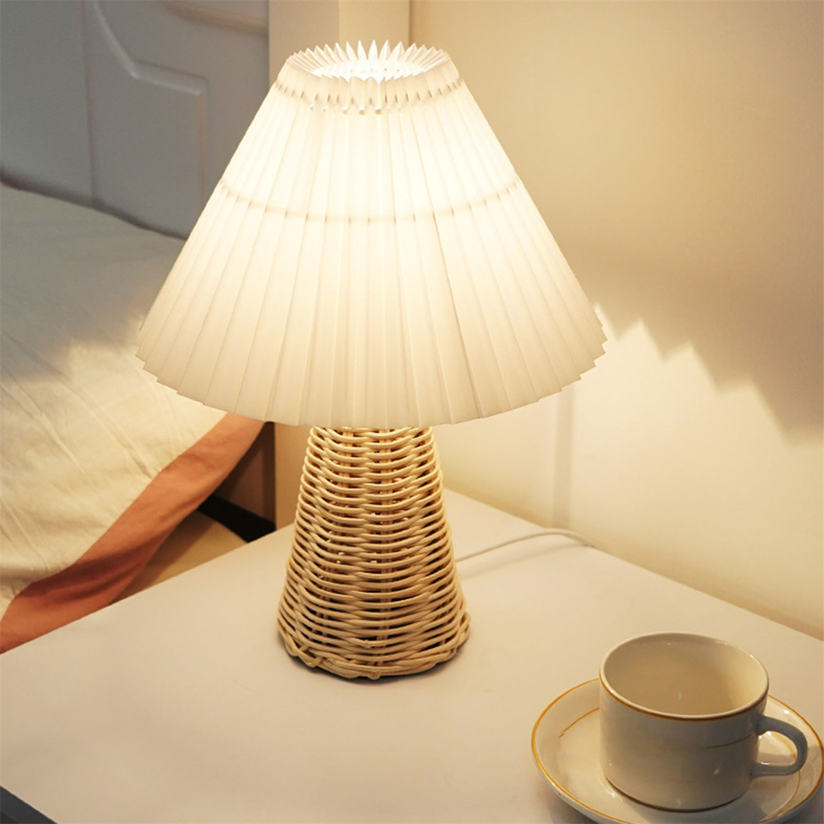 Eine Lampe mit geflochtenem Fuß und plissiertem Schirm, neben einer Tasse auf dem Tisch.
