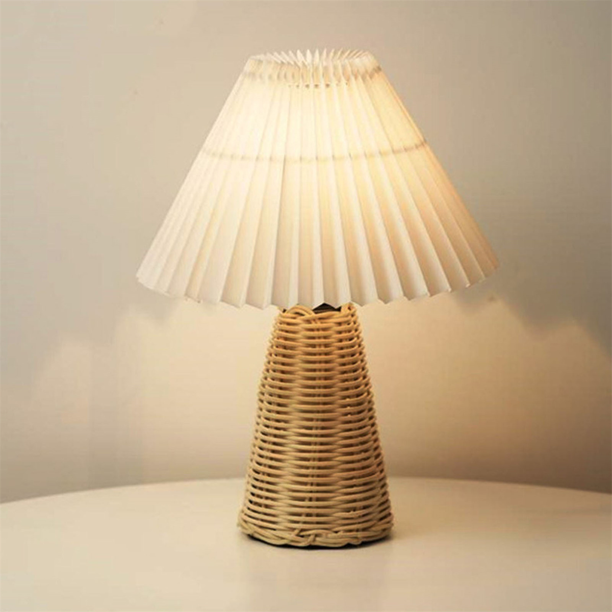 Tischlampe mit gewebtem Sockel und plissiertem Lampenschirm auf einem weißen Tisch.