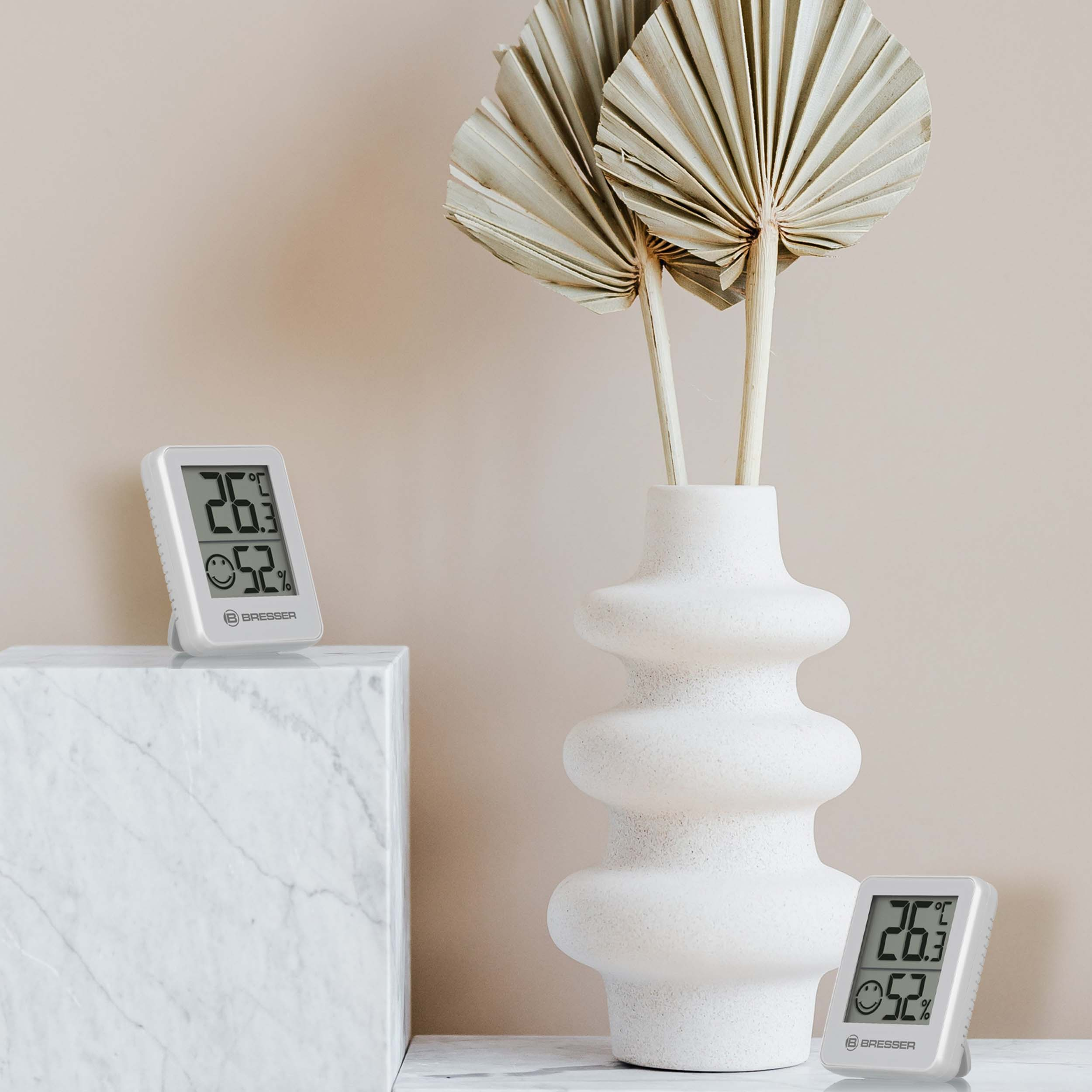 Zwei weiße Digitalthermometer mit Messwerten und eine Vase mit getrockneten Blättern auf beigem Hintergrund.