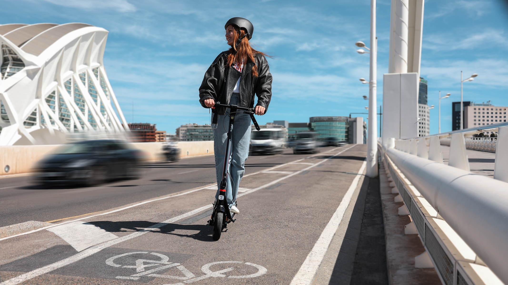 Eine Frau fährt mit einem Elektroroller auf einer Brücke. Stadtgebäude und Autos im Hintergrund.