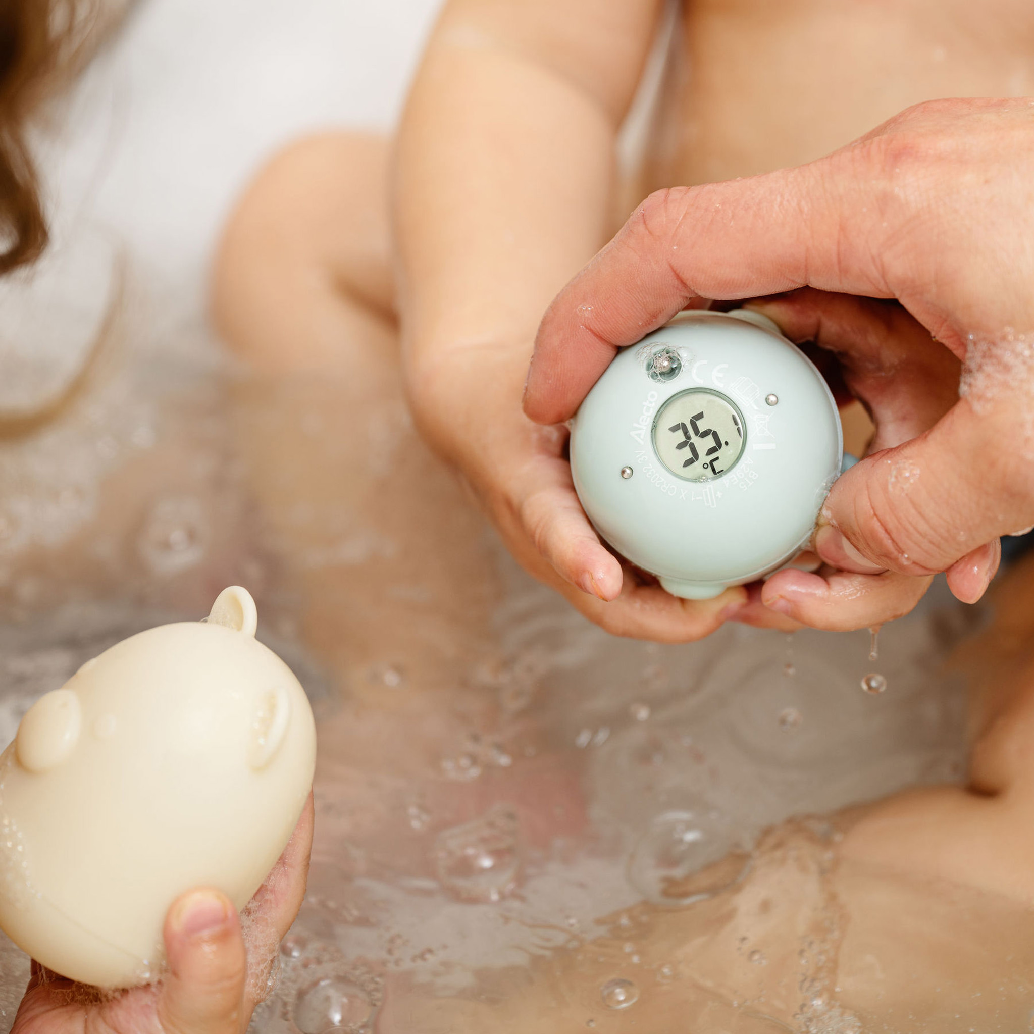 Ein Baby und die Hände einer Person halten ein blaues digitales Badethermometer. Blasen im Wasser.