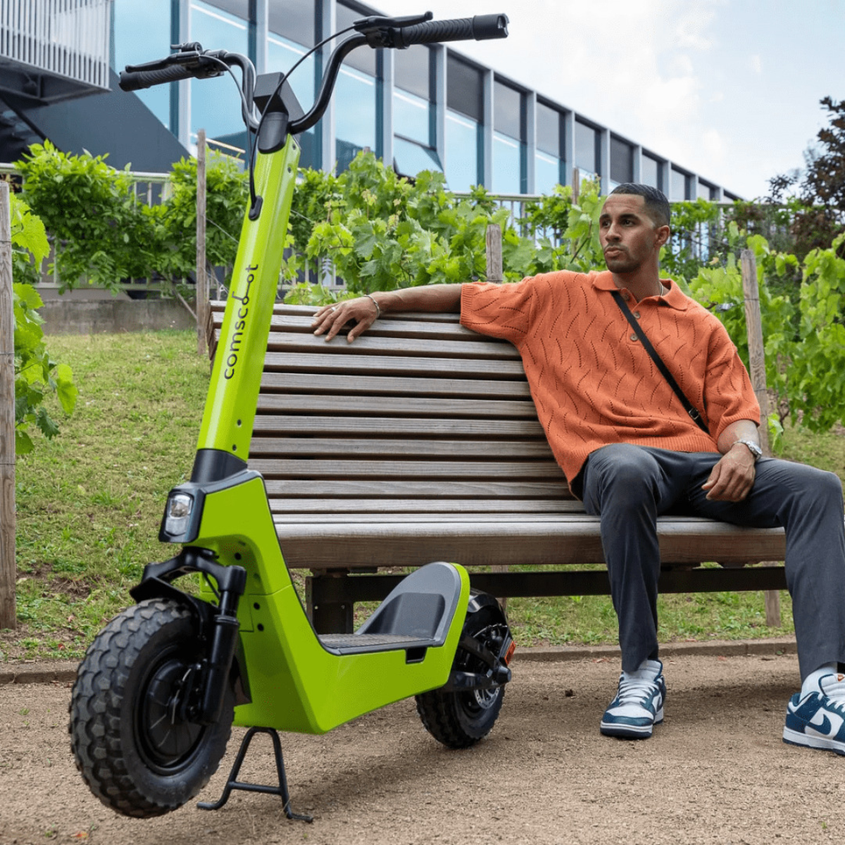 Ein Mann sitzt auf einer Bank neben einem grünen Roller. Er trägt ein orangefarbenes Hemd.