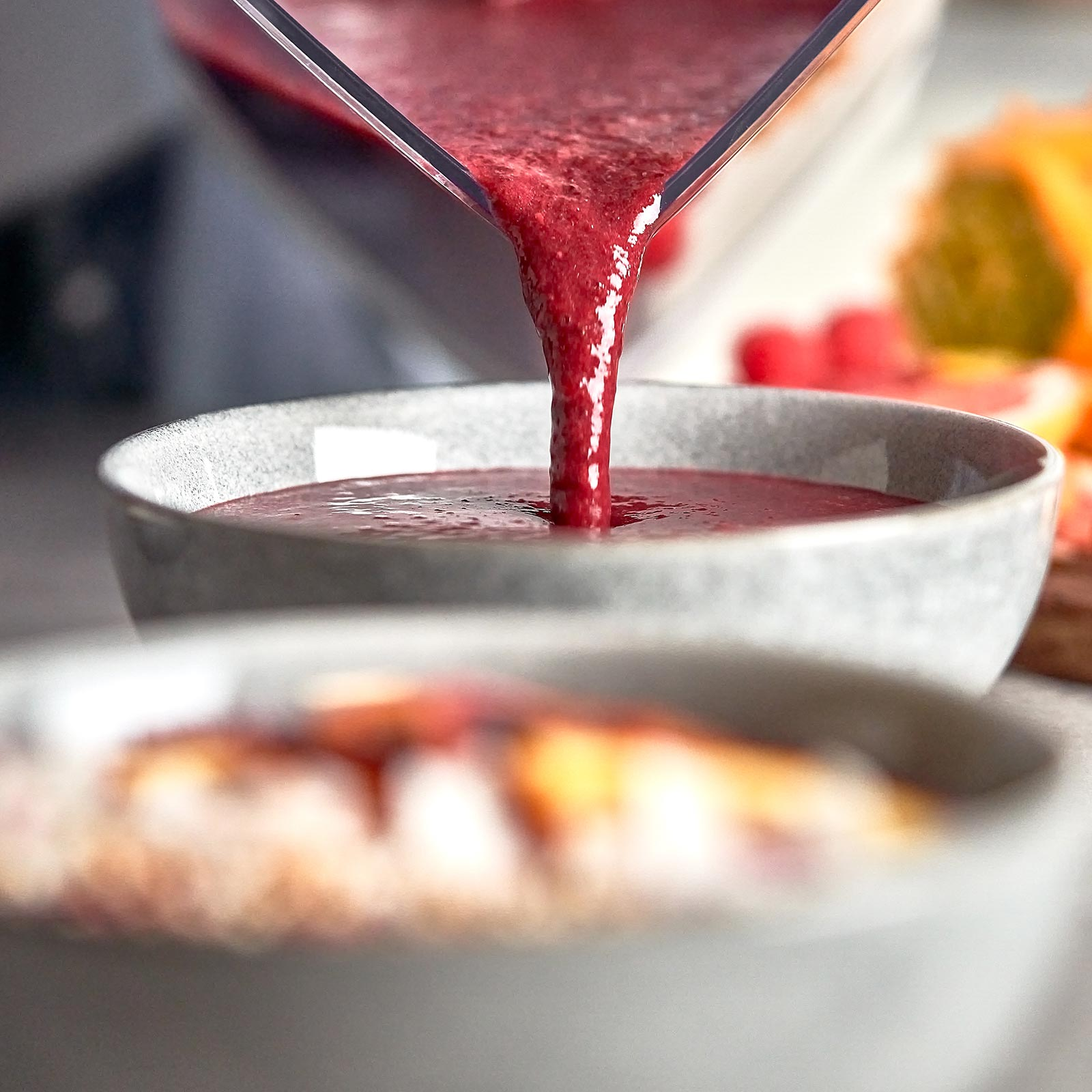 Roter Smoothie wird aus einem Mixer in eine graue Schüssel gegossen. Andere Schüsseln und Früchte im Hintergrund.