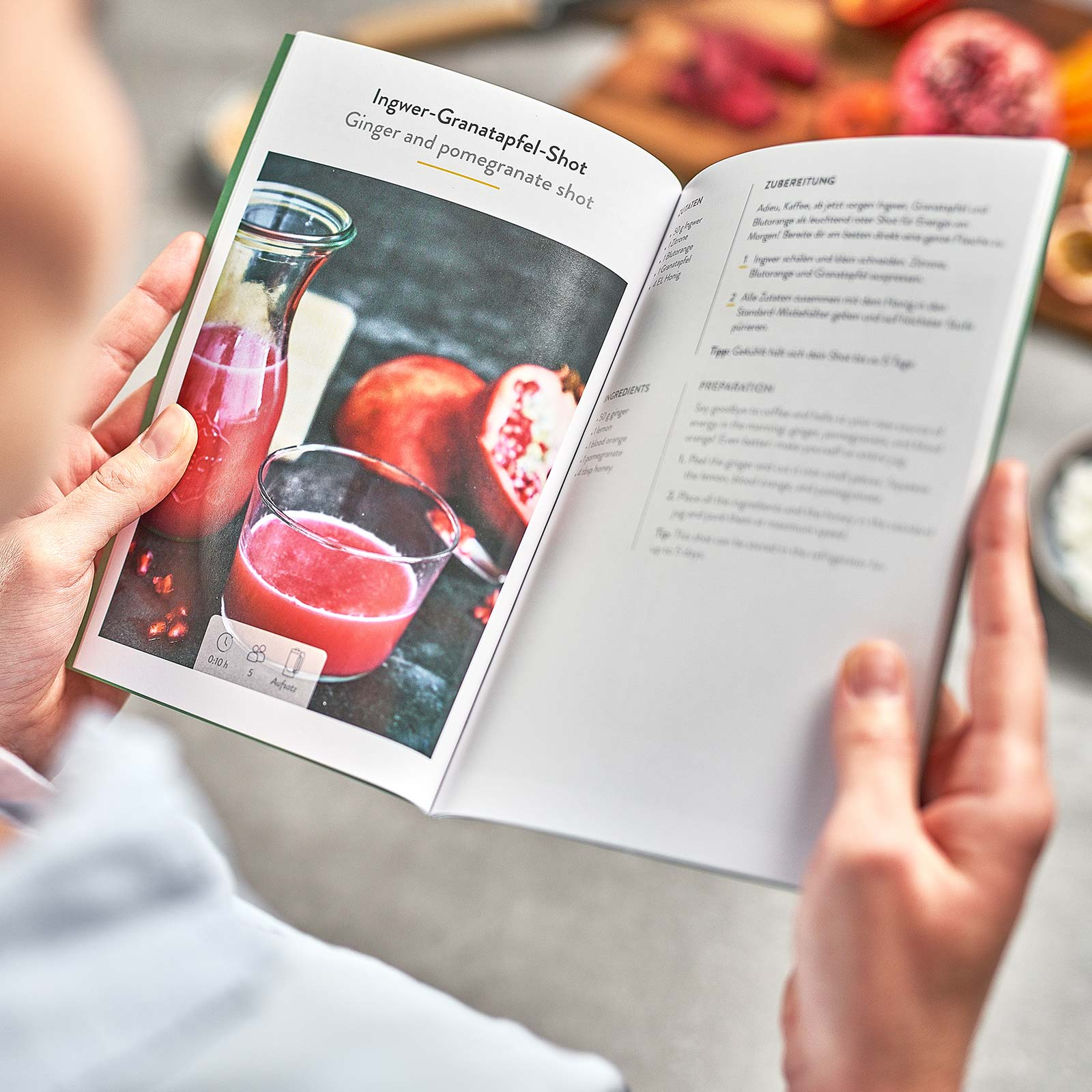Offenes Buch mit Saft- und Granatapfel-Shot-Rezept, gehalten von Händen.