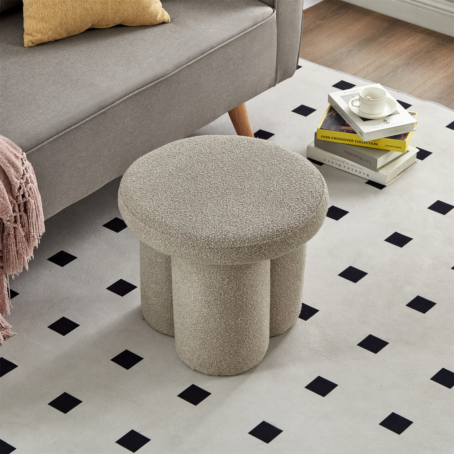 Ein dreibeiniger beiger Hocker steht vor einem grauen Sofa auf einem Teppich mit schwarzen Quadratmustern.