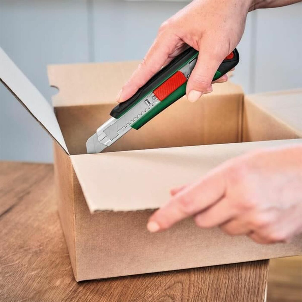 Eine Person schneidet mit einem Teppichmesser einen Karton auf einem Holztisch auf.