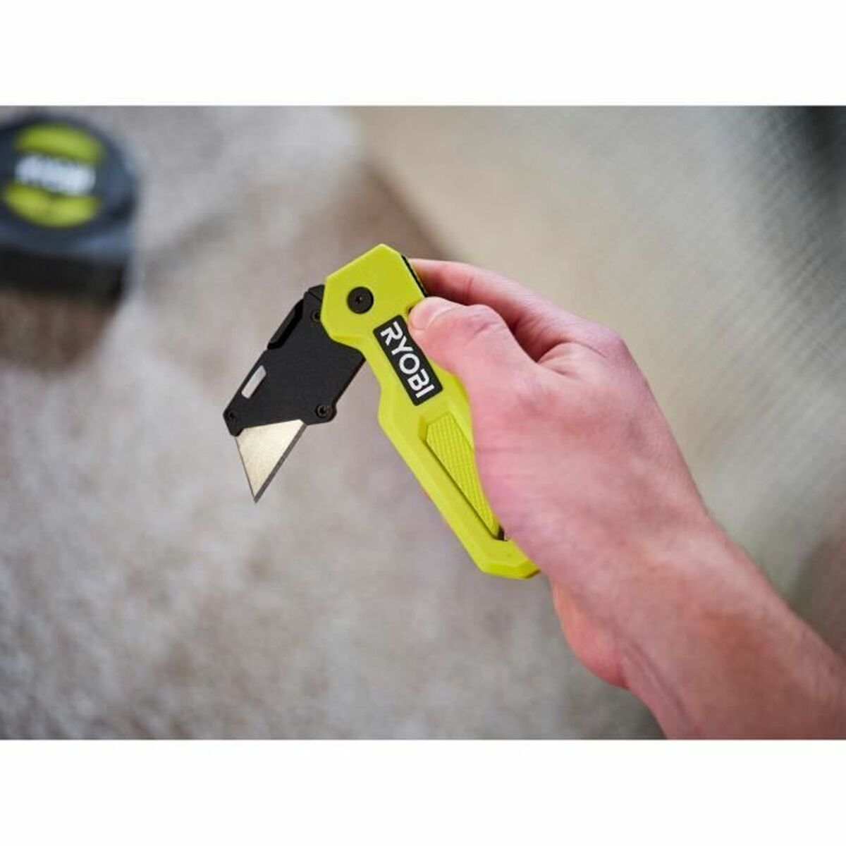 Eine Hand, die ein gelb-schwarzes Universalmesser hält. Ein Maßband ist im Hintergrund.