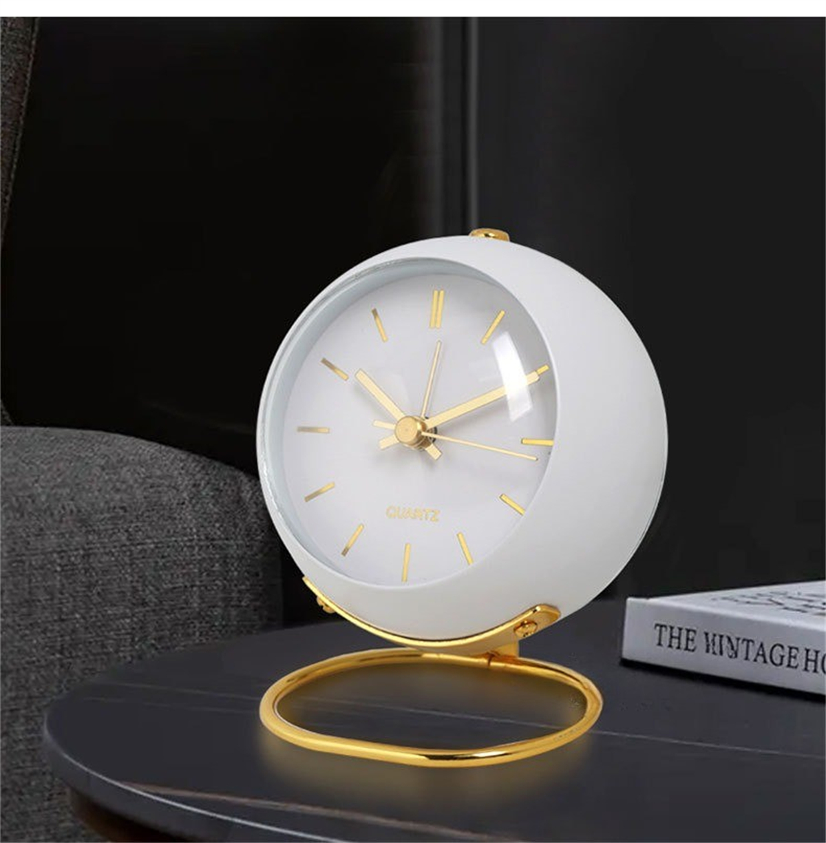 Eine weiße, kugelförmige Uhr mit goldenen Akzenten, auf einem schwarzen Tisch. Das Zifferblatt hat goldene Zeiger und Zahlen. Buch im Hintergrund.