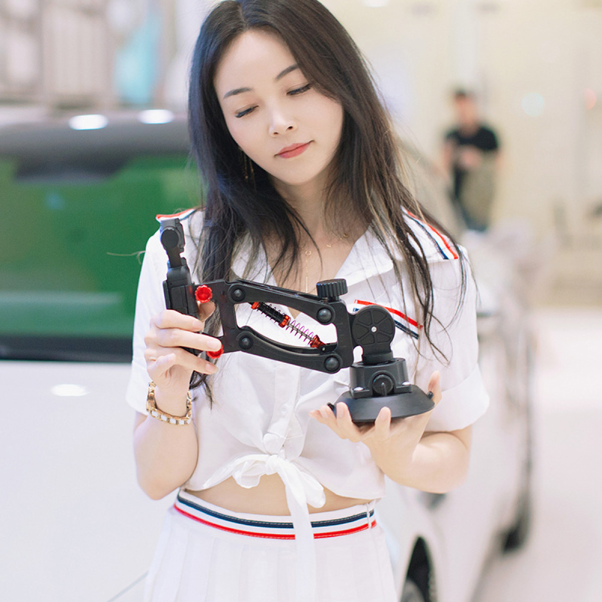 Kobieta w białym stroju trzymająca czarne narzędzie, rozmyte tło z samochodem.