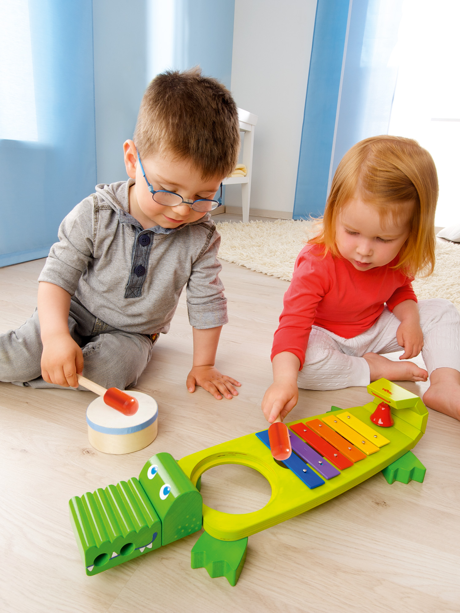 Zwei Kinder spielen mit Spielzeug, eines mit einer Trommel, das andere mit einem Xylophon. Holzboden.