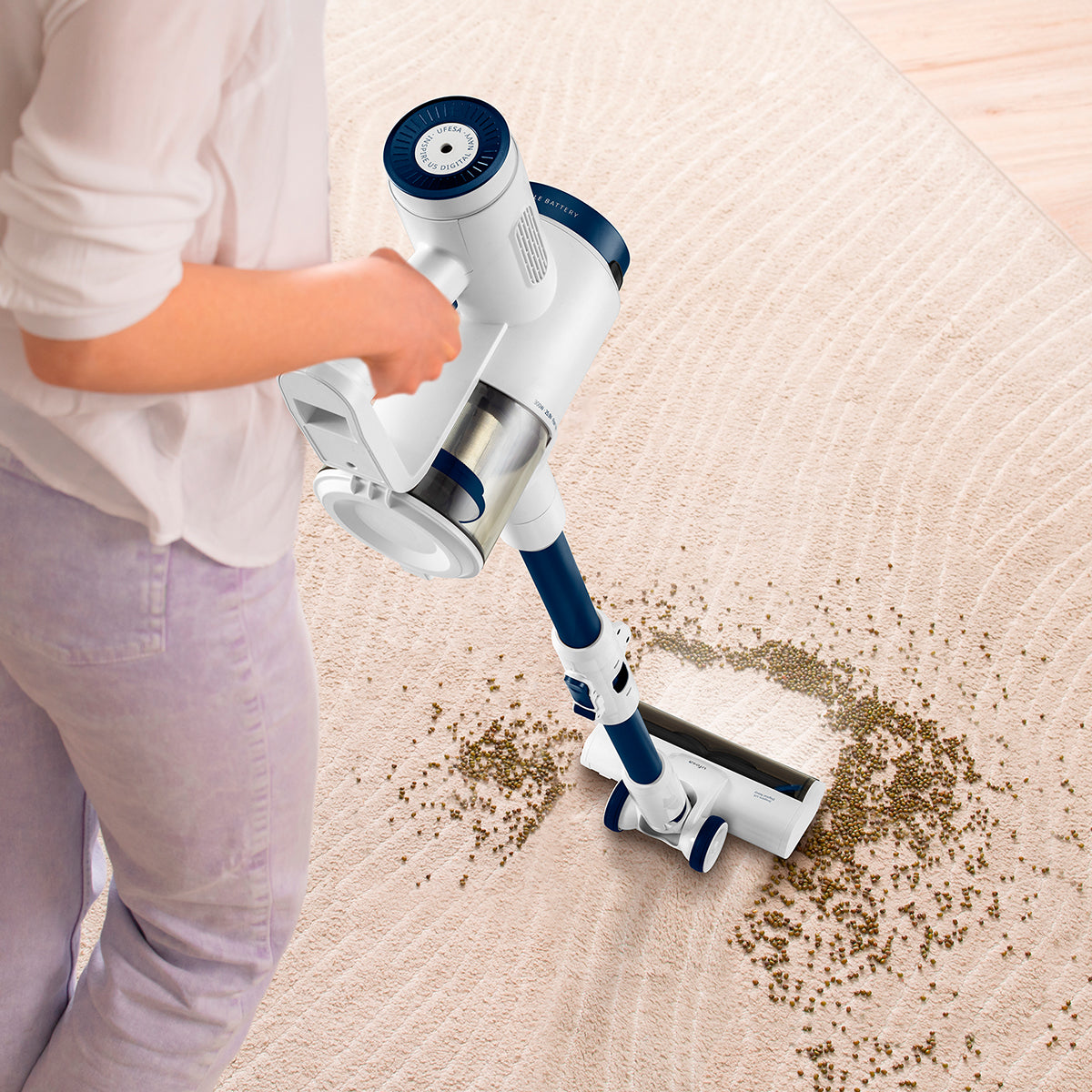 Persona aspirando una alfombra beige con una aspiradora de varilla blanca y azul.