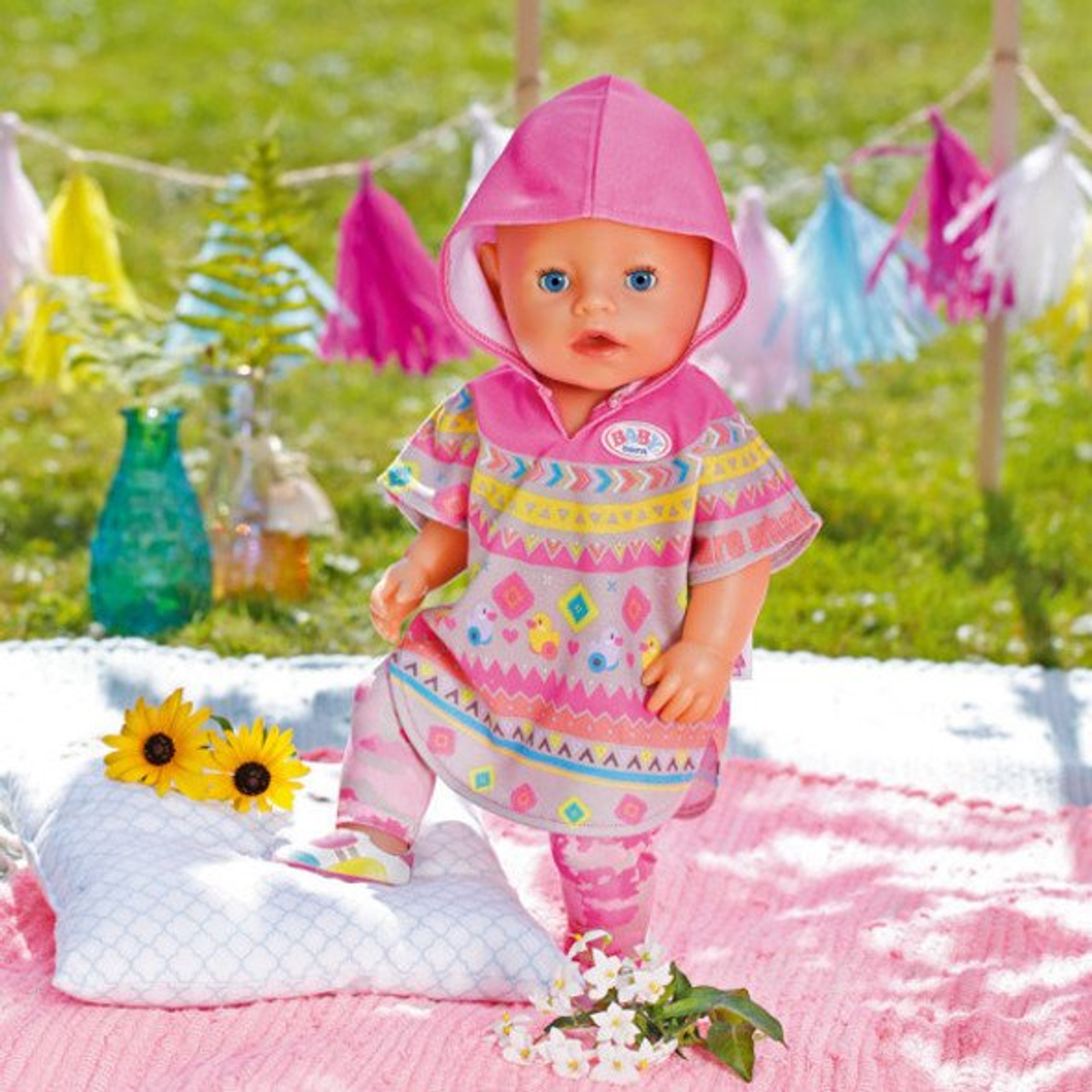 Eine Puppe in einem rosa Kapuzenponcho und Leggings steht auf einer rosa Oberfläche, mit Blumen und Dekorationen.