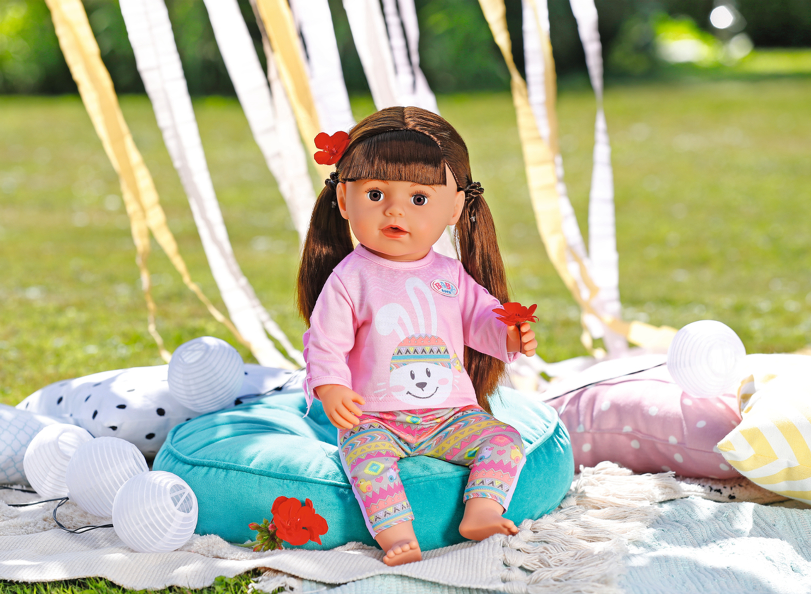 Puppe mit rosa Hemd und bunter Hose, sitzt draußen auf blauem Kissen, sonnige Umgebung.