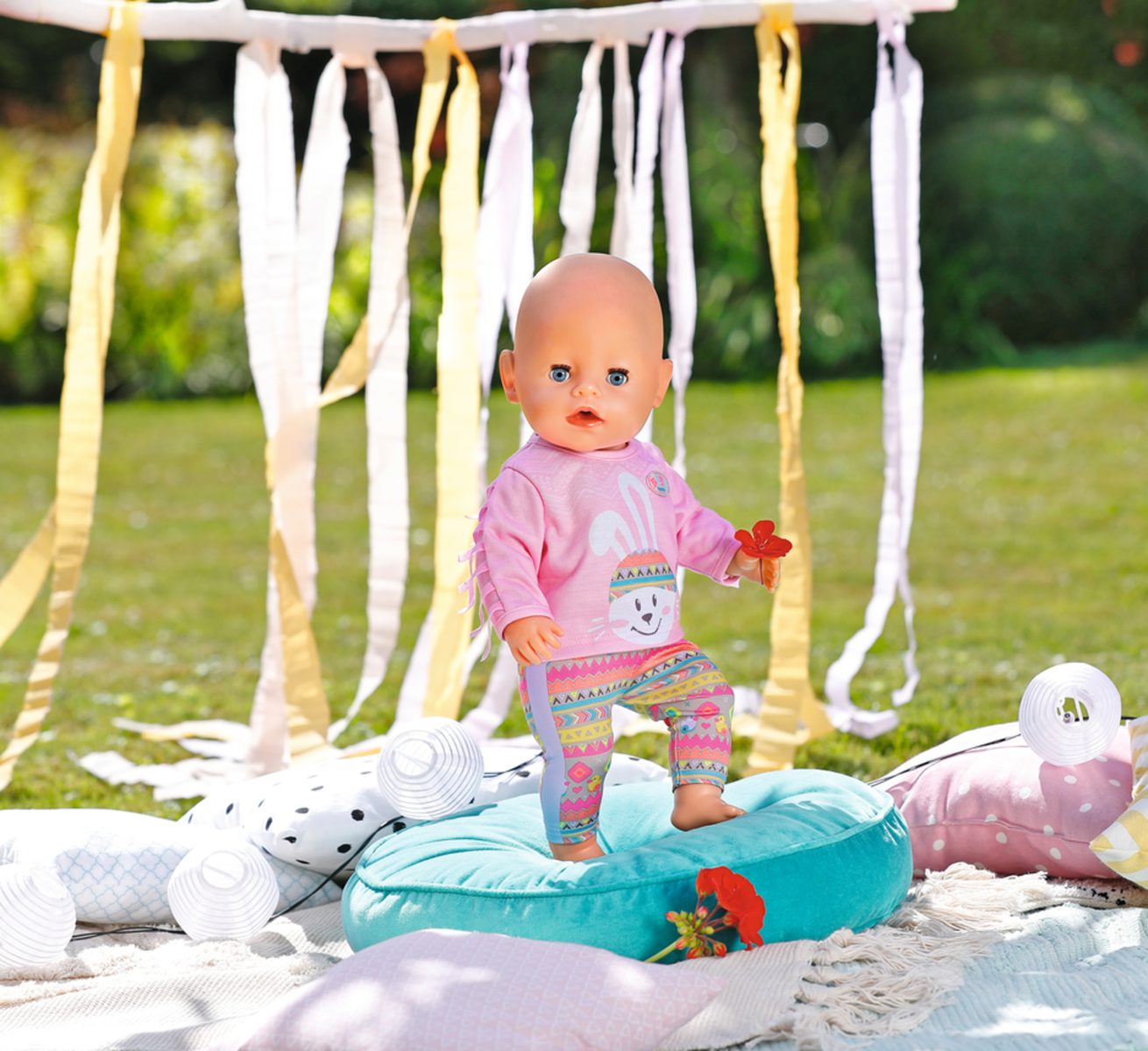 Eine Babypuppe in rosa Kleidung, steht auf einem türkisfarbenen Kissen im Garten.