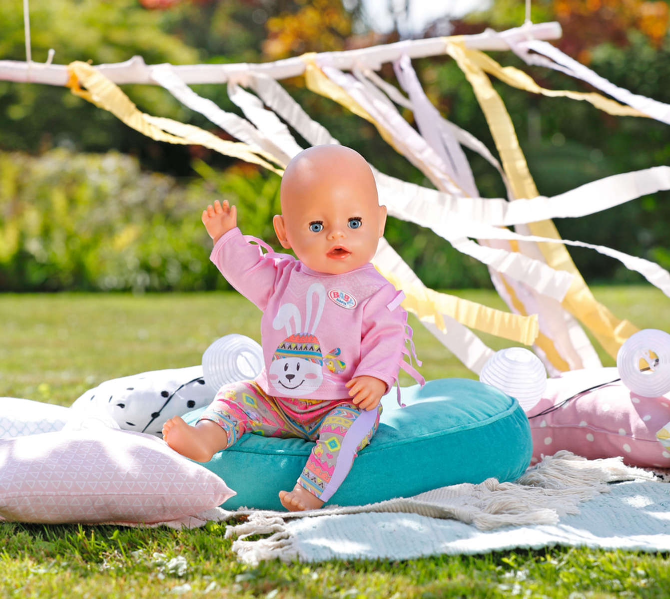 Eine Babypuppe in rosa Kleidung sitzt auf einem türkisfarbenen Kissen auf Gras, mit dekorativen Bändern.