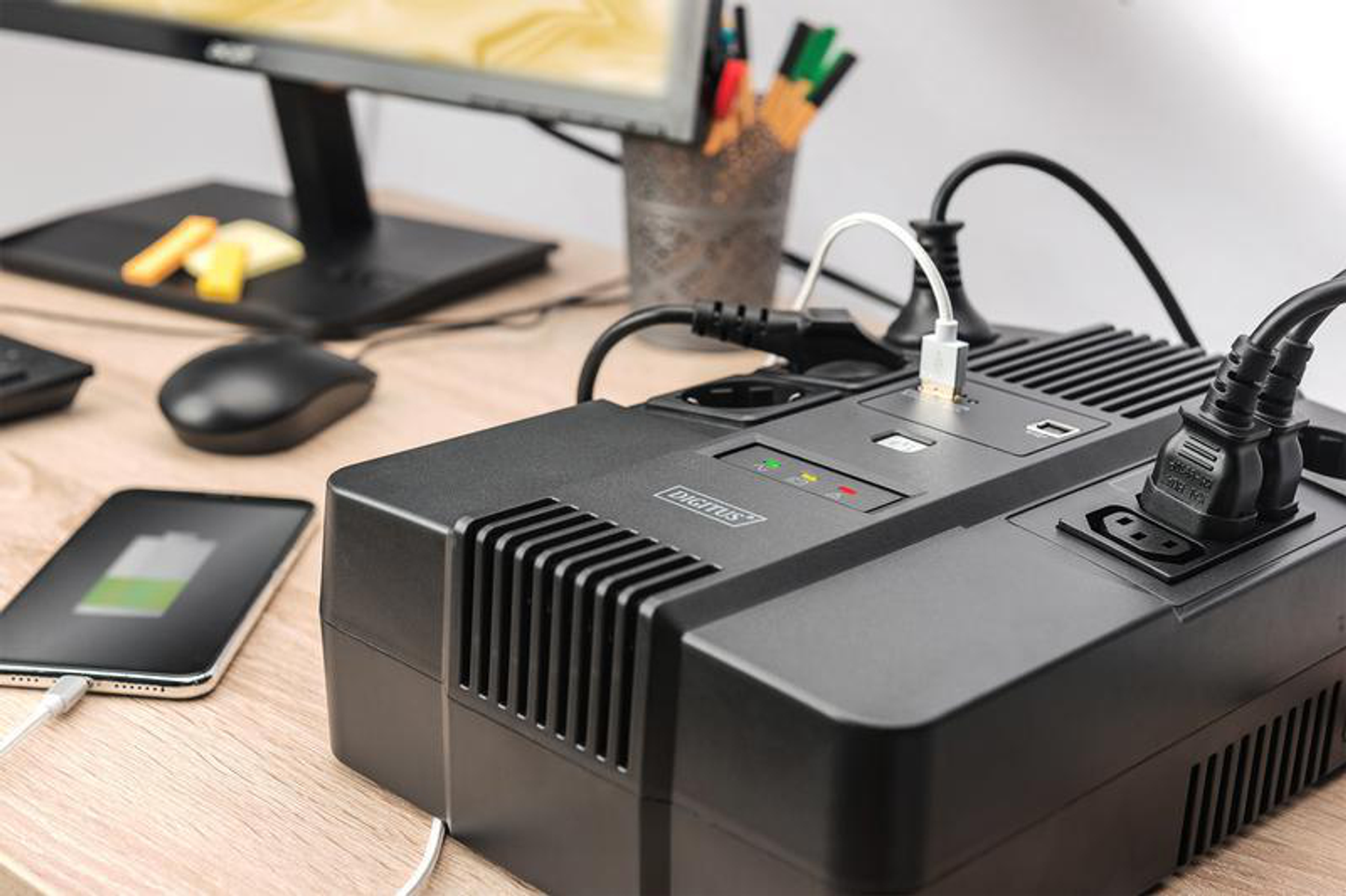 Eine schwarze USV mit Steckern, ein ladendes Telefon und ein Computermonitor auf einem Holztisch.
