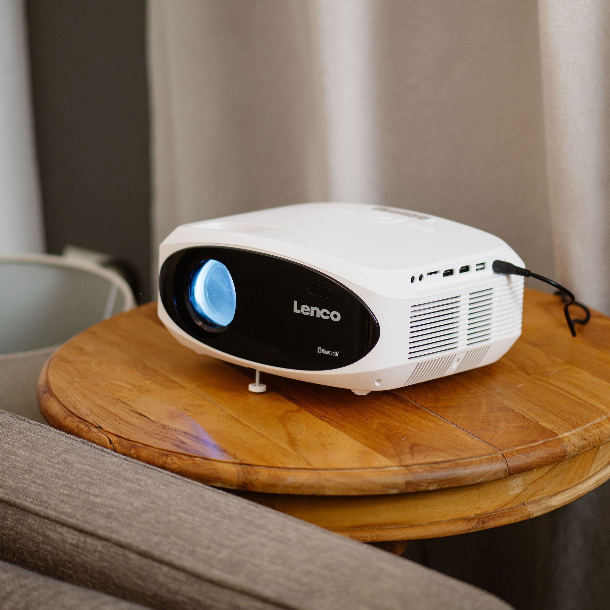 Biały projektor Lenco na drewnianym stole. Szara kanapa w tle.