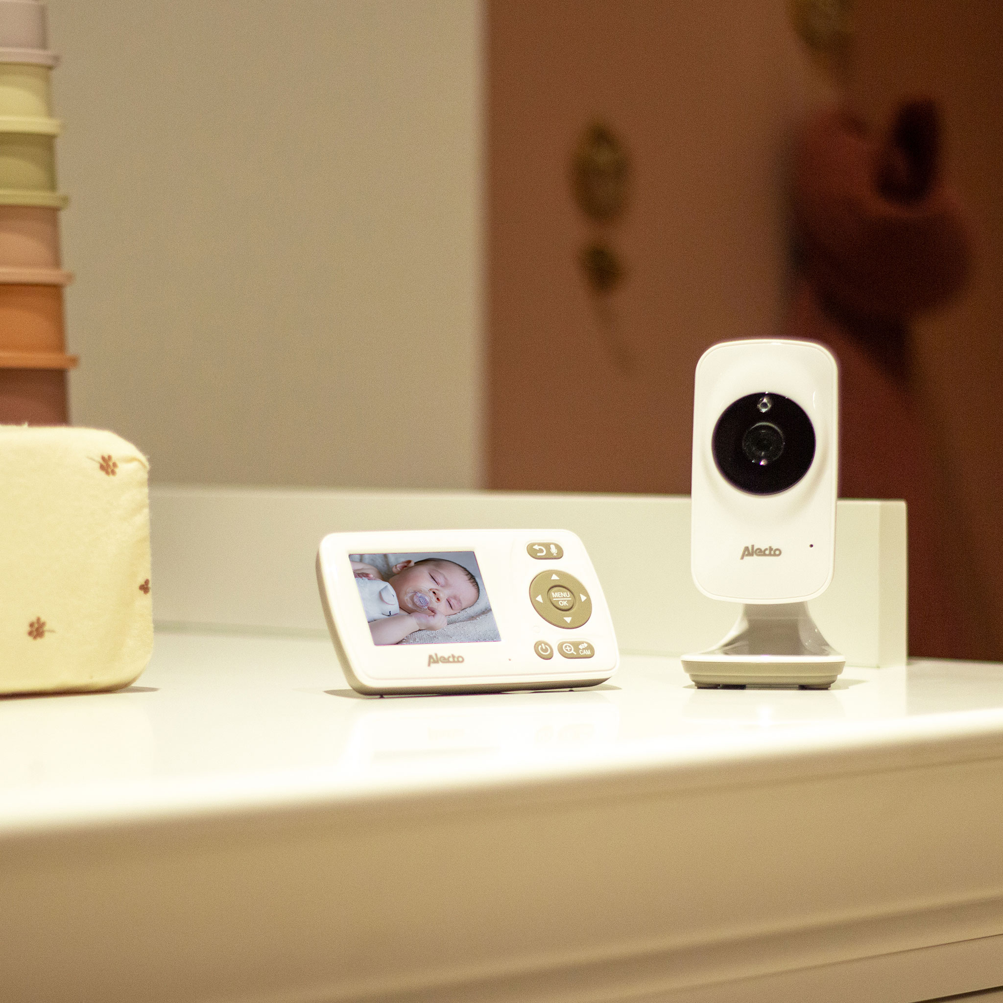 Baby monitor. Monitor displays a baby. Camera is white, placed on a table. Pink background.