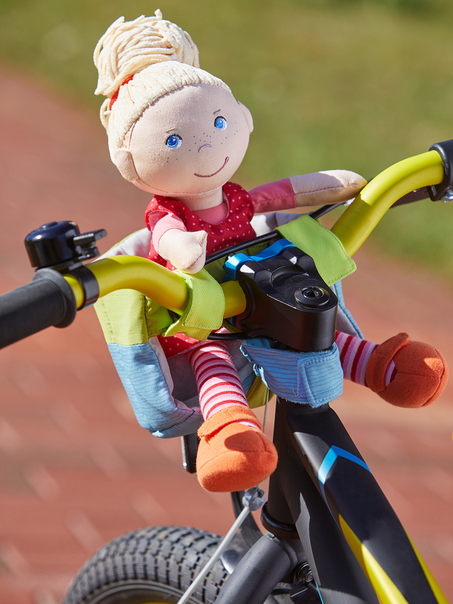 Puppe auf einem Fahrrad. Puppe trägt rotes Kleid, blaue Augen, blondes Haar. Fahrrad ist gelb und schwarz.