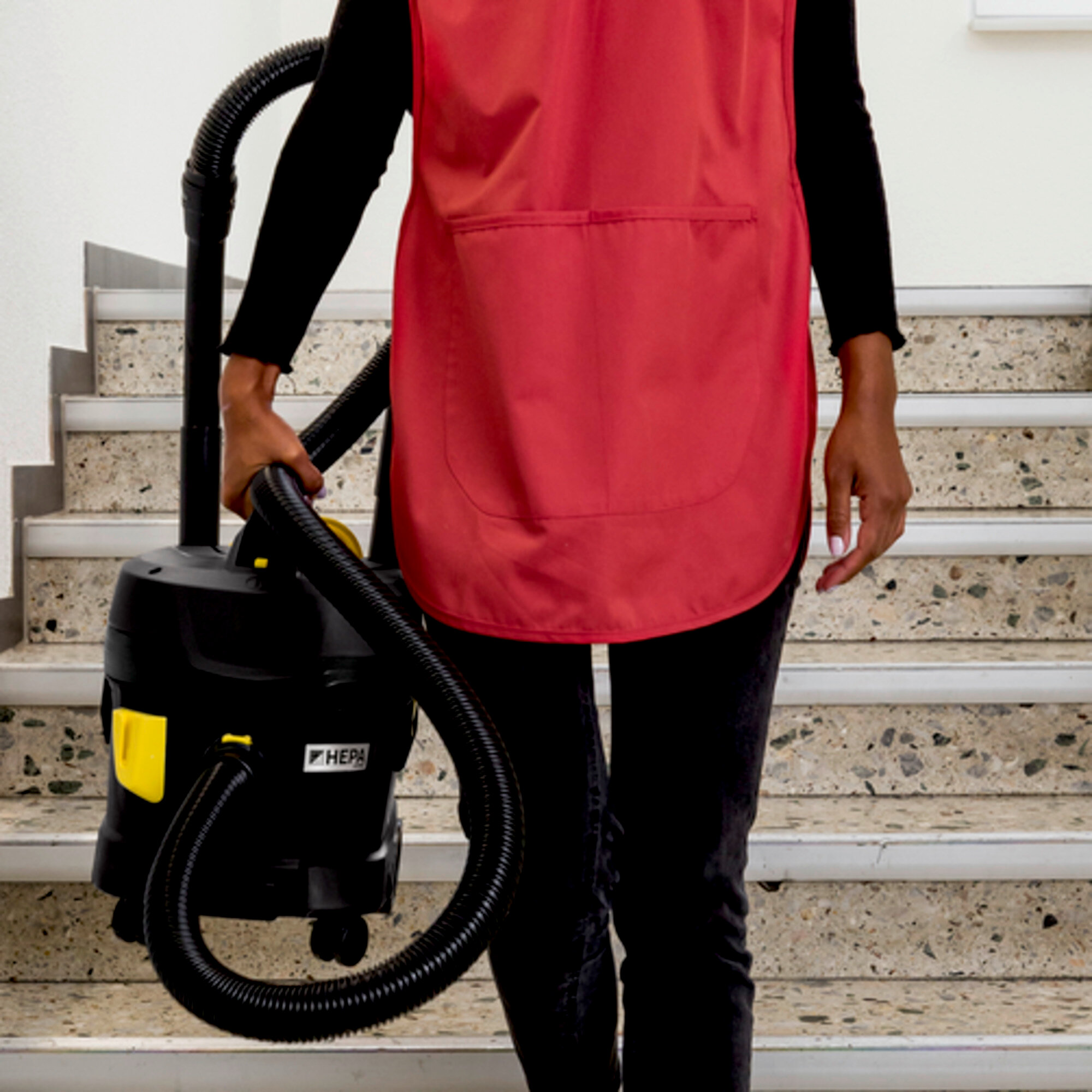 Osoba w czerwonym fartuchu trzymająca czarny odkurzacz na schodach.