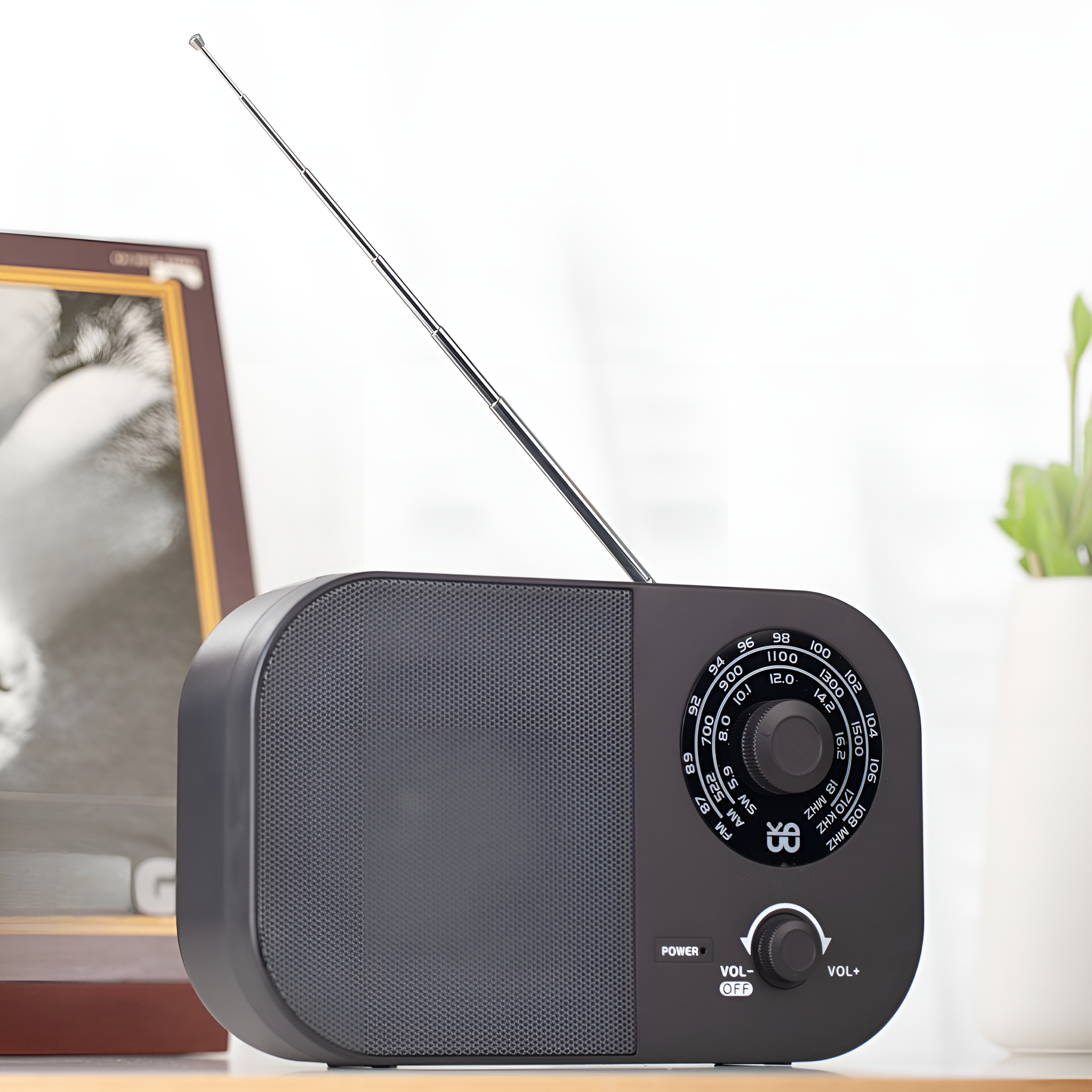 Schwarzes Radio auf einer Holzoberfläche, mit Drehknopf, Lautsprecher und Antenne. Ein Bilderrahmen im Hintergrund.
