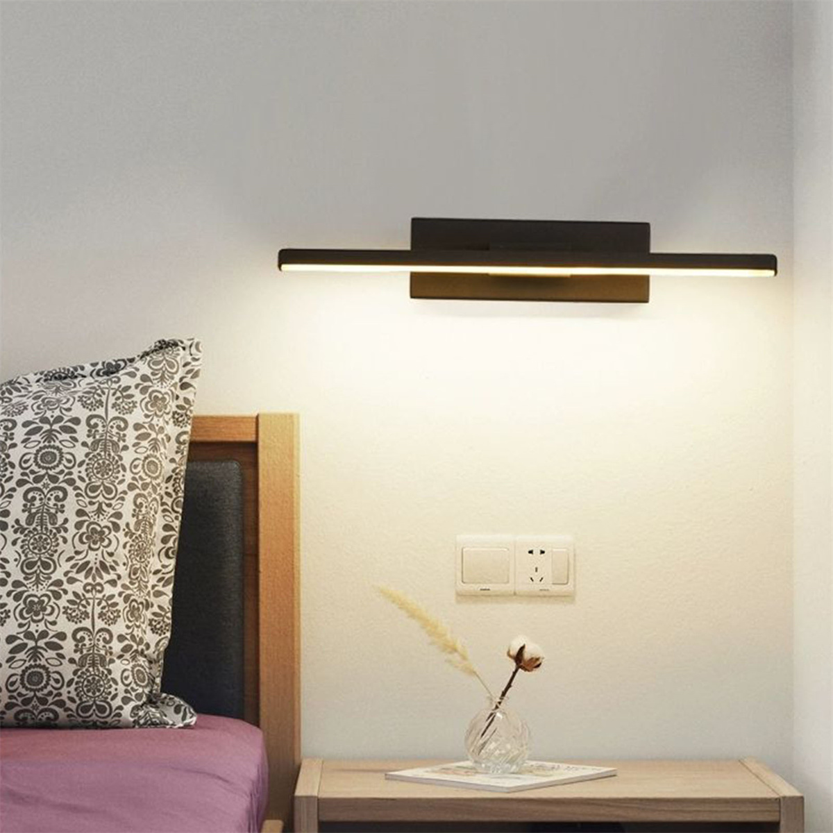 Eine Wandleuchte in Schwarz mit horizontaler Lichtleiste, mit Bett und Vase im Blickfeld.