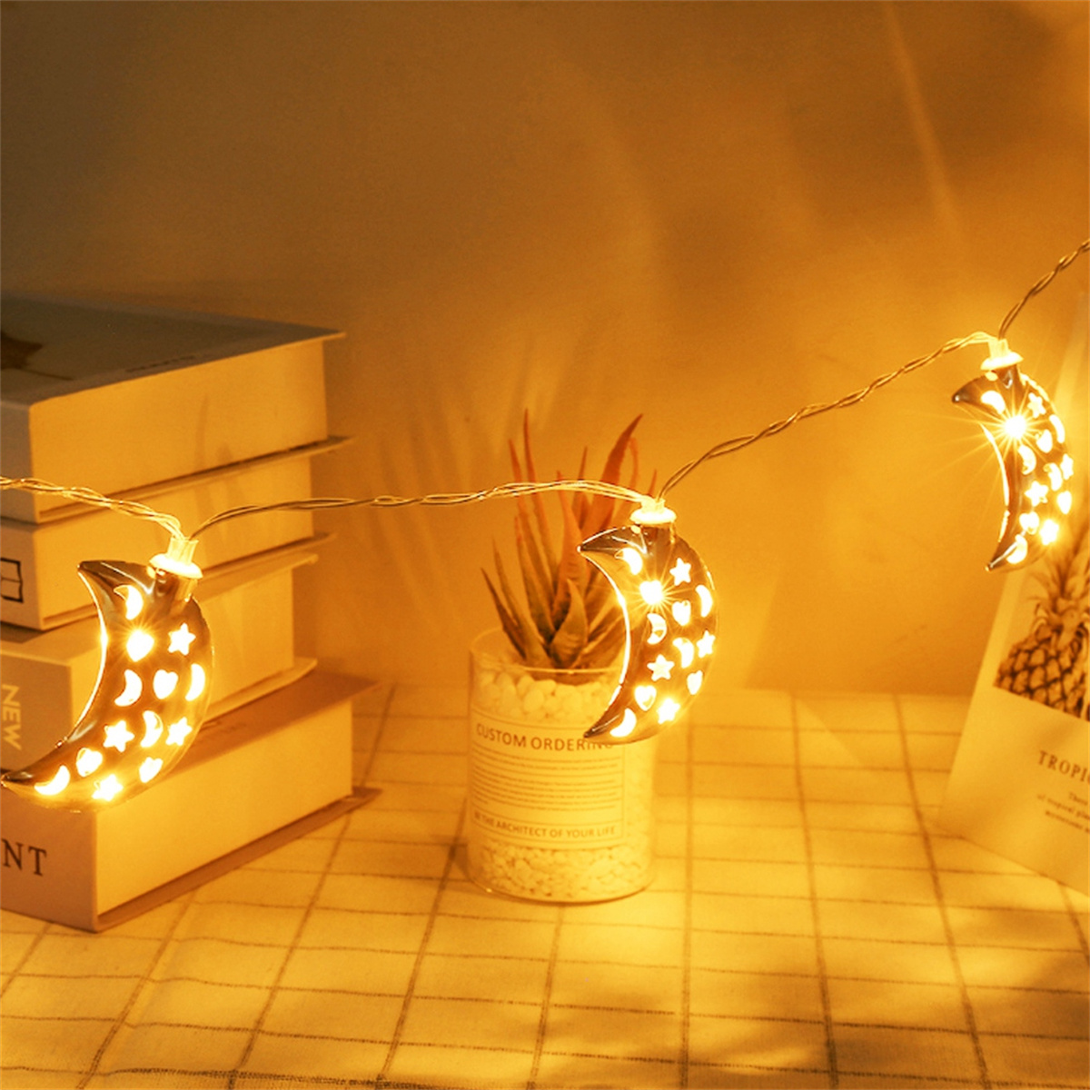 Lichterkette mit Halbmond-Dekorationen und warmem Schein, vor einem Hintergrund mit Büchern und Pflanzen.