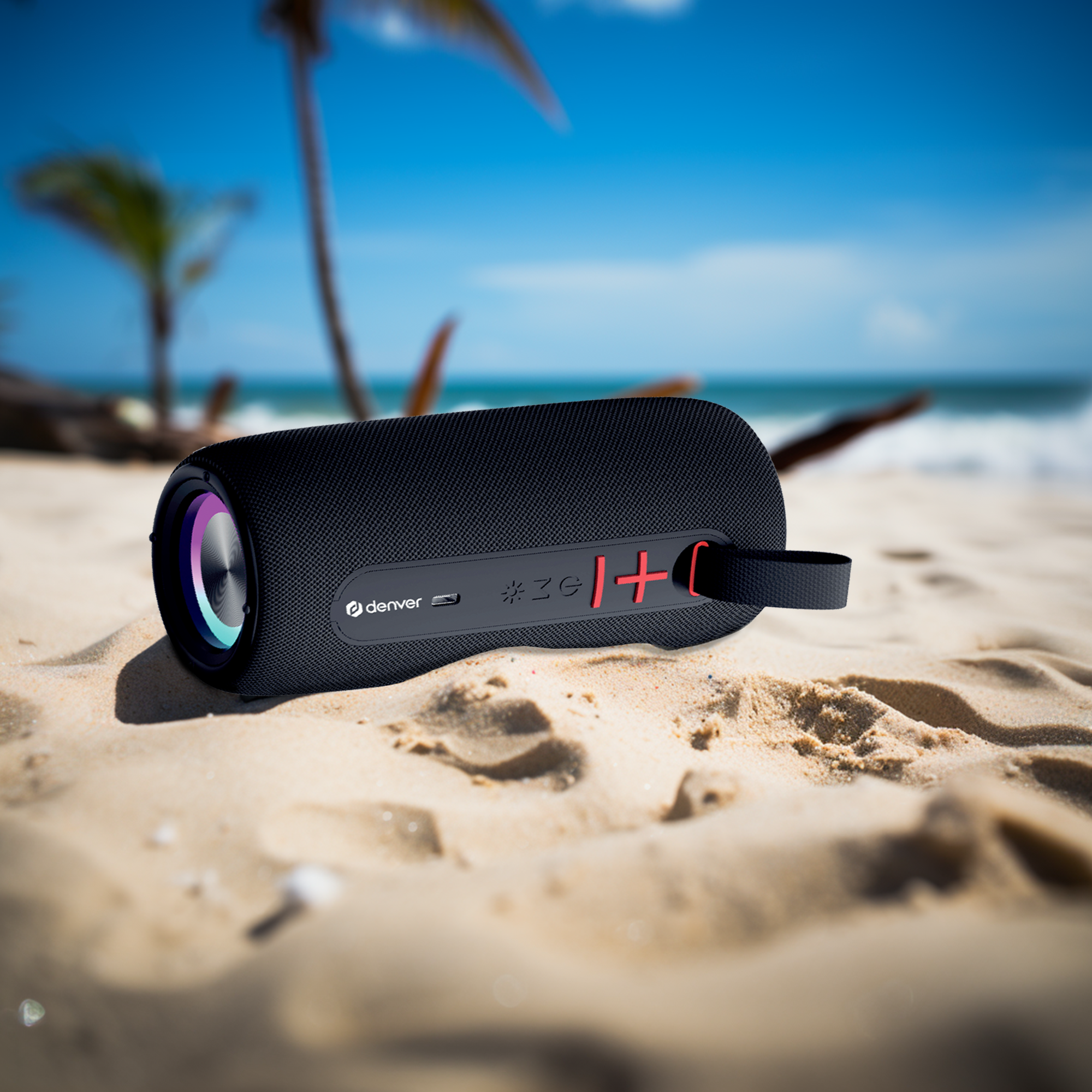 Czarny głośnik Denver na piaszczystej plaży z palmami i błękitnym morzem.