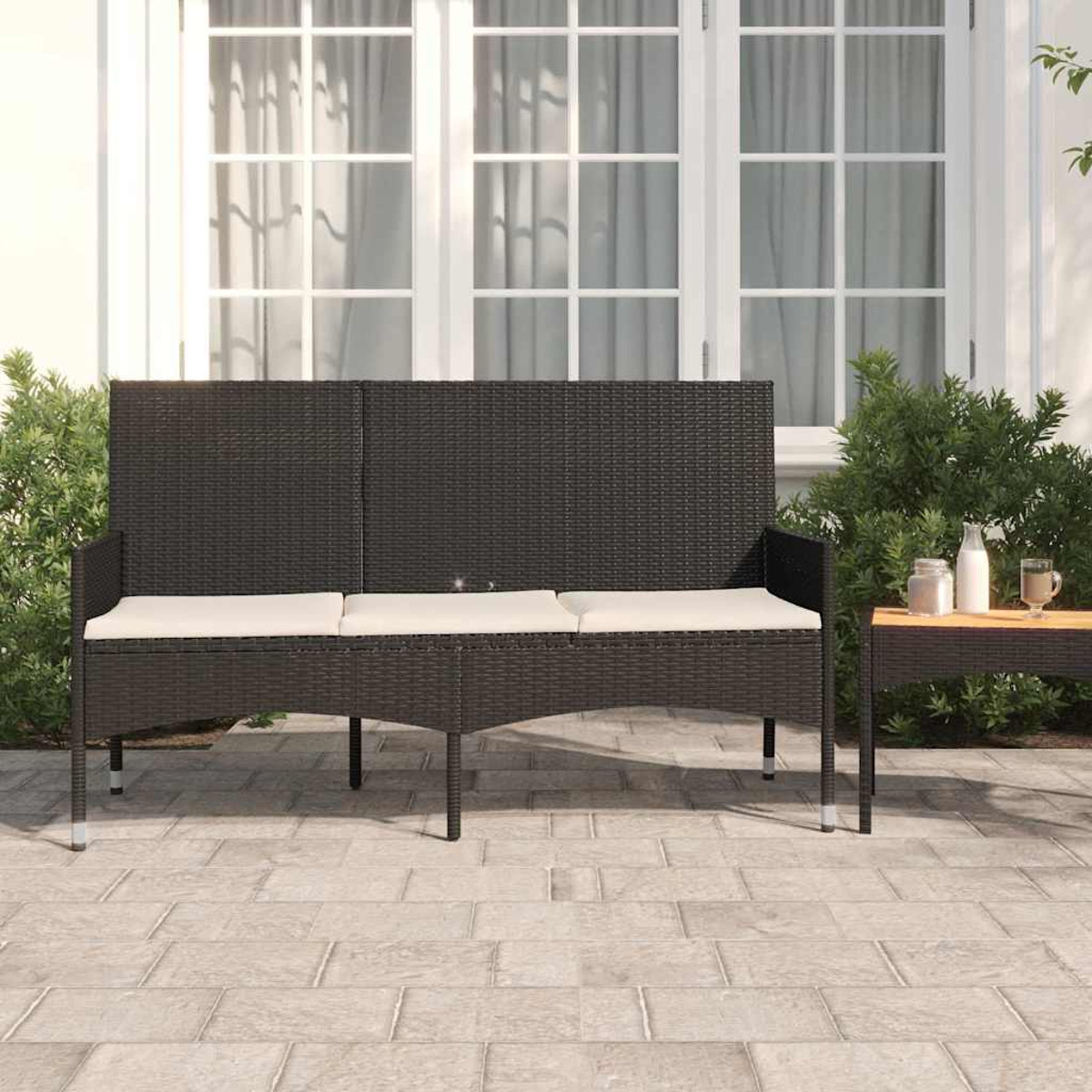 Schwarzes Rattansofa mit cremefarbenen Kissen auf einer Terrasse mit Tisch. Büsche und ein Gebäude im Hintergrund.