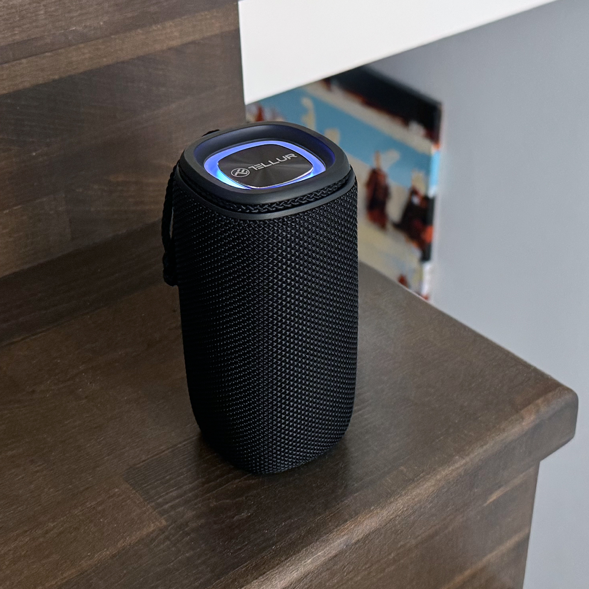 Czarny, cylindryczny głośnik z niebieskim podświetlanym wierzchem stoi na drewnianych schodach.