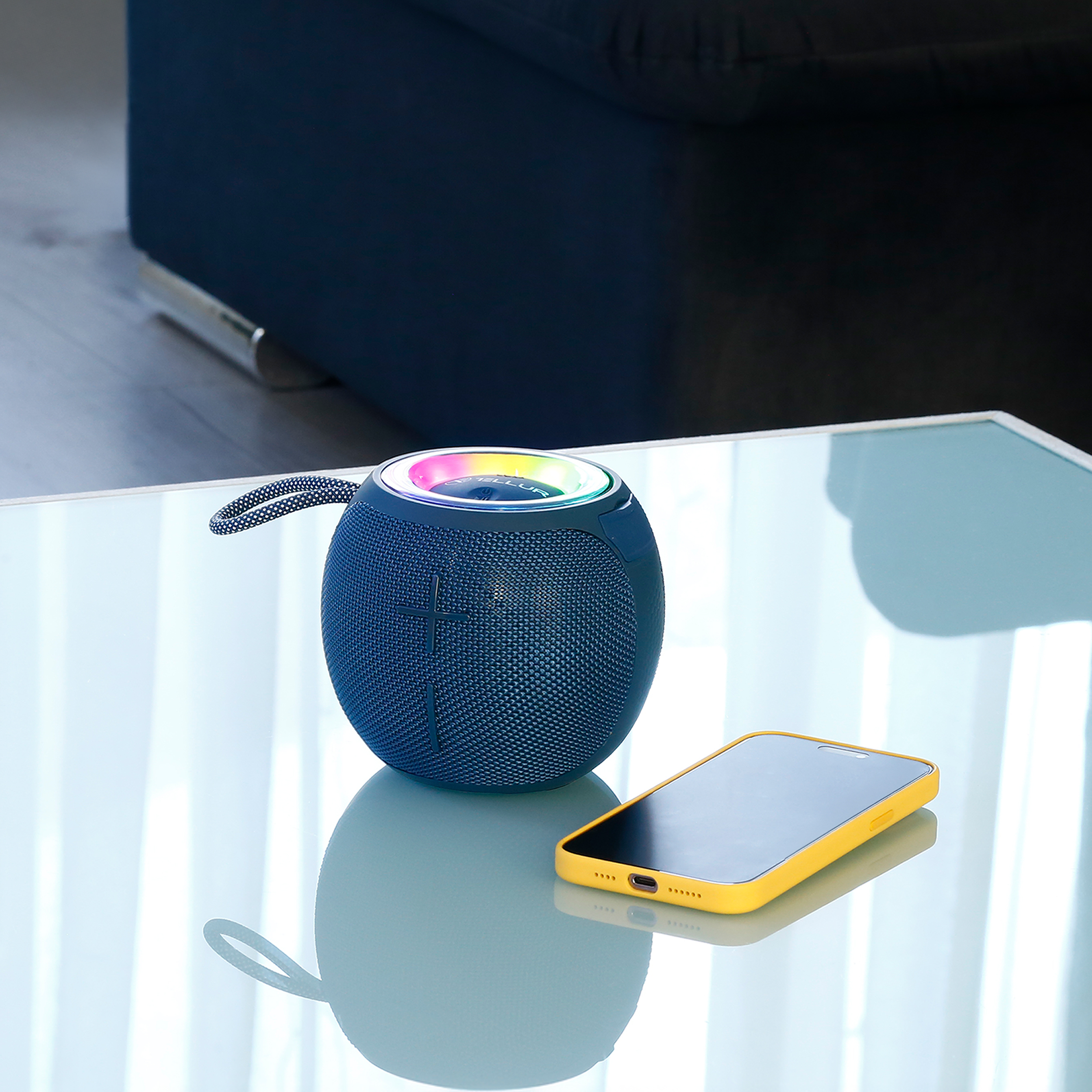 Niebieski głośnik z telefonem na szklanym stole. Czarne tło kanapy.