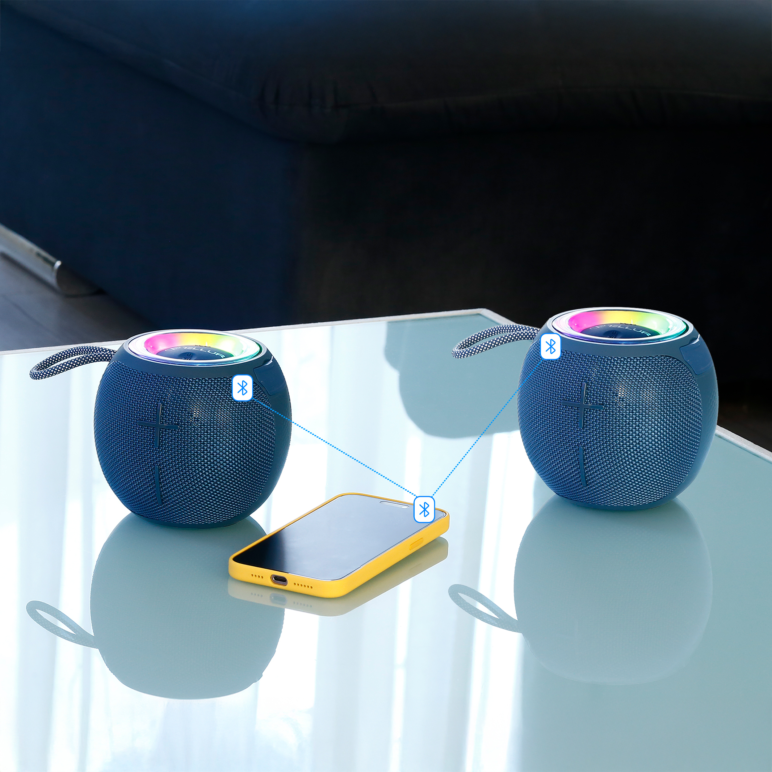 Dwa niebieskie głośniki połączone z telefonem na szklanym stole. Czarne tło kanapy.