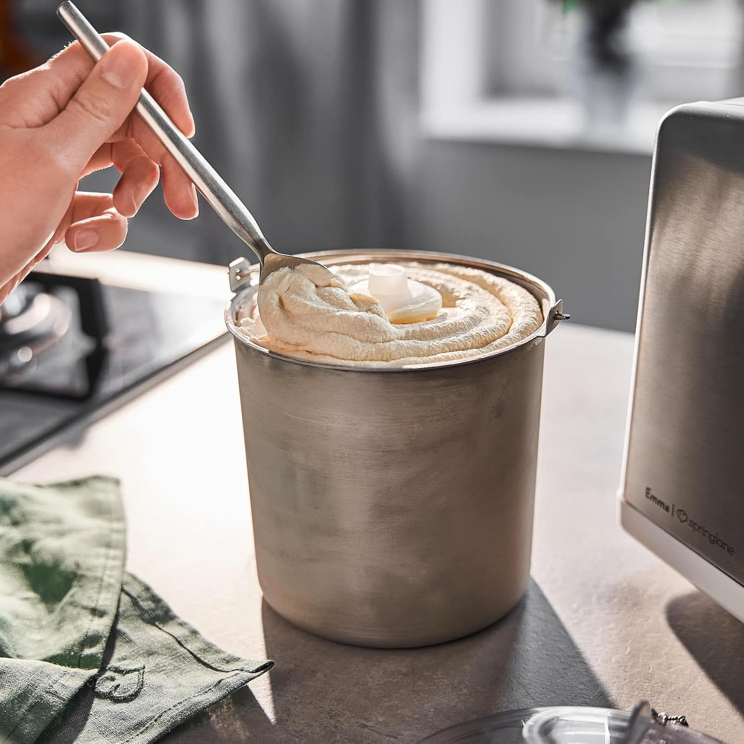 Eine Hand schöpft Eiscreme aus einem Metallbehälter mit weißer Oberfläche auf einer Arbeitsplatte mit grünem Tuch.