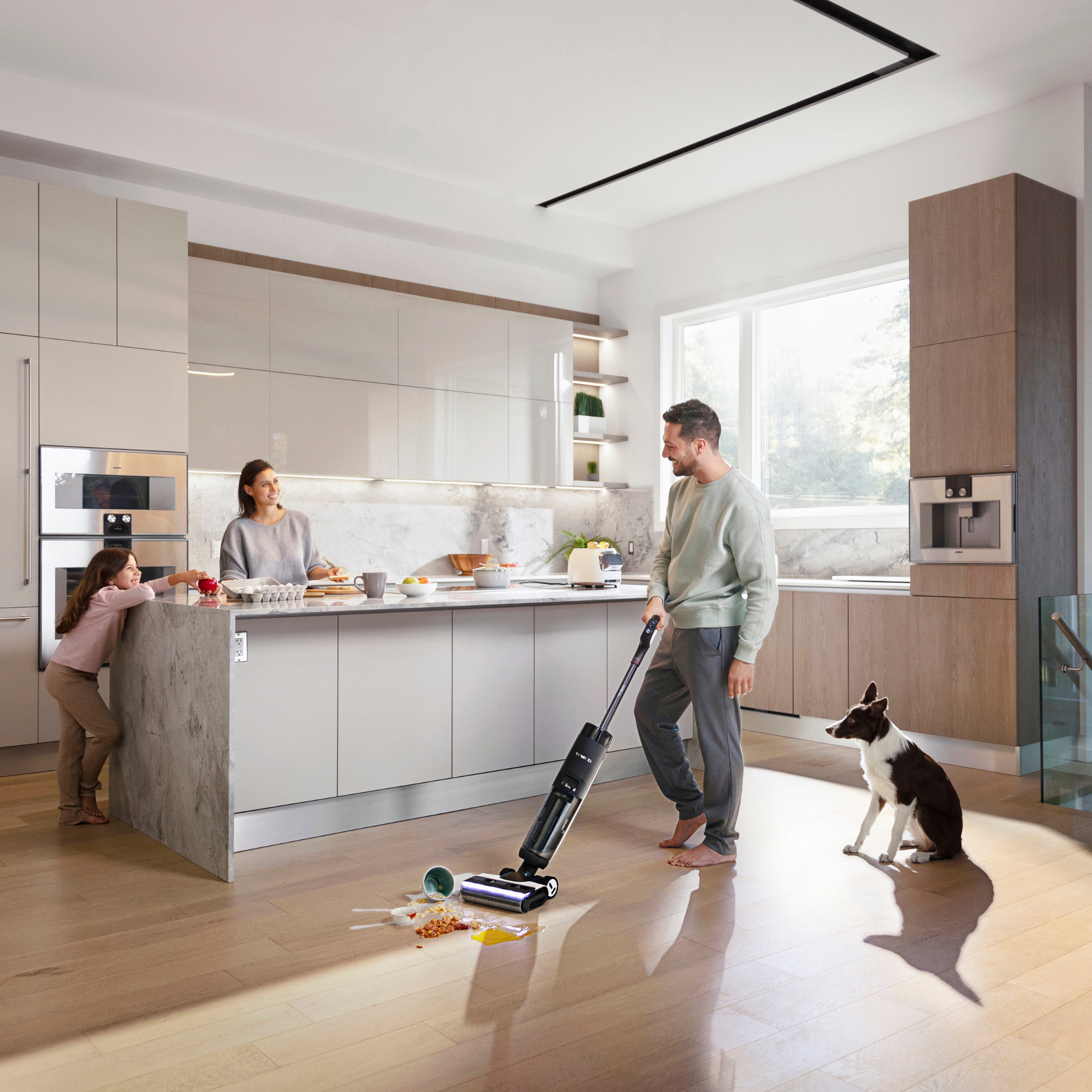 Una famiglia in una cucina moderna. Un uomo aspira, una donna cucina, una bambina e un cane sono presenti.