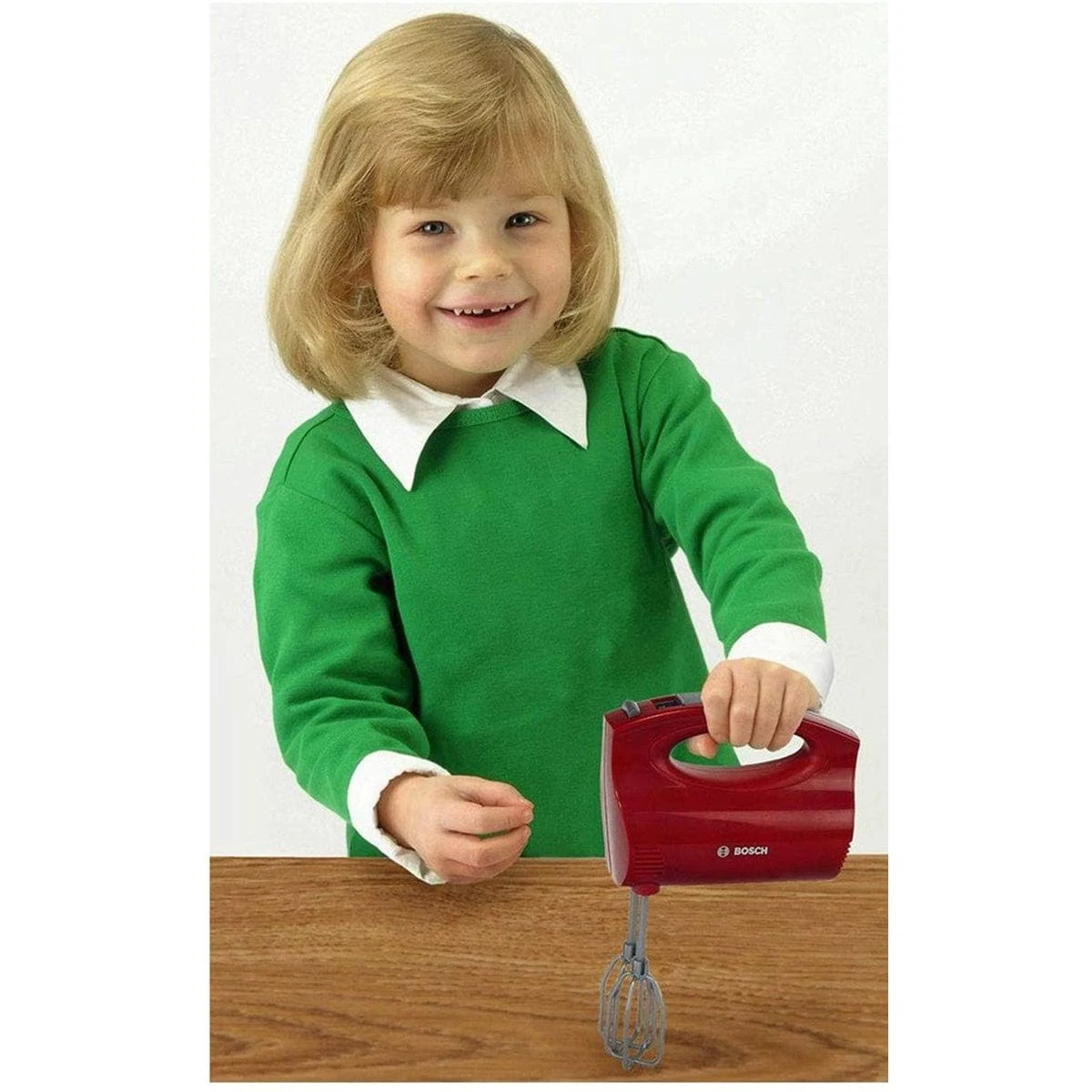 Niña rubia con camisa verde sosteniendo una batidora roja. Sonriendo a la cámara.