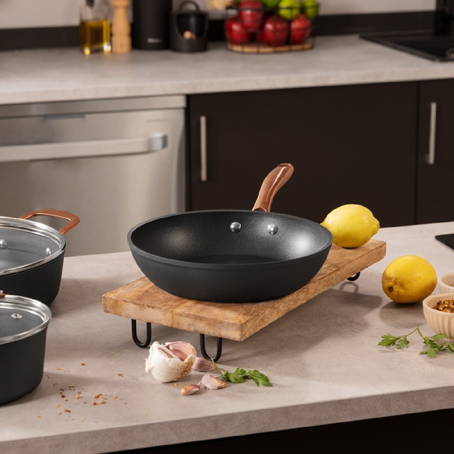 Escena de cocina con sartén negra en soporte de madera, limones y ajo en encimera.