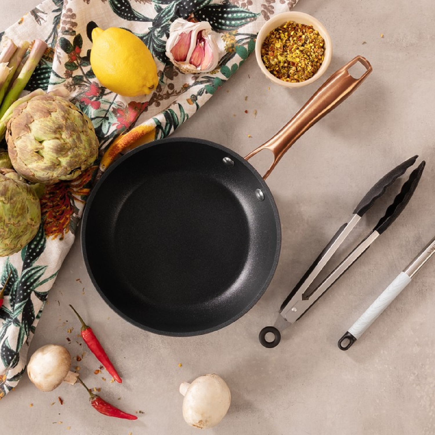 Una sartén con ingredientes como alcachofas, limón, ajo y pimientos rojos. También hay pinzas y especias.