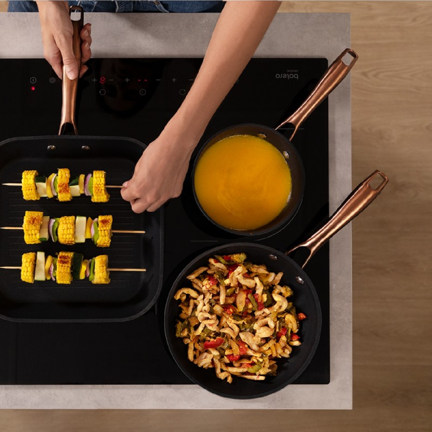 Widok z góry na czarną płytę kuchenną z jedzeniem w patelniach i na szaszłykach, obsługiwanych przez osobę.