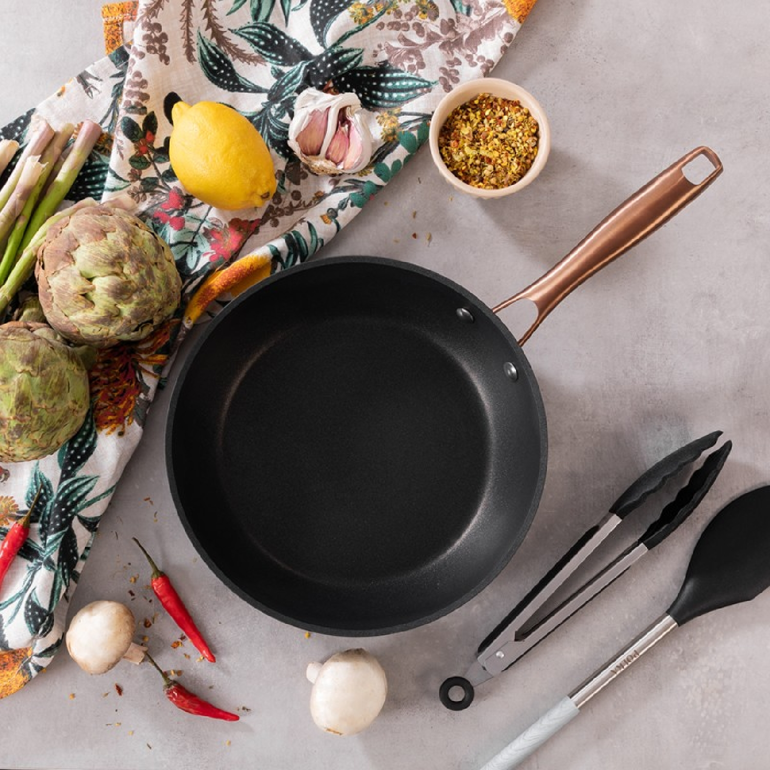 Patelnia, karczochy, czosnek, cytryna, papryczki chili, szczypce i szpatułka na stole.