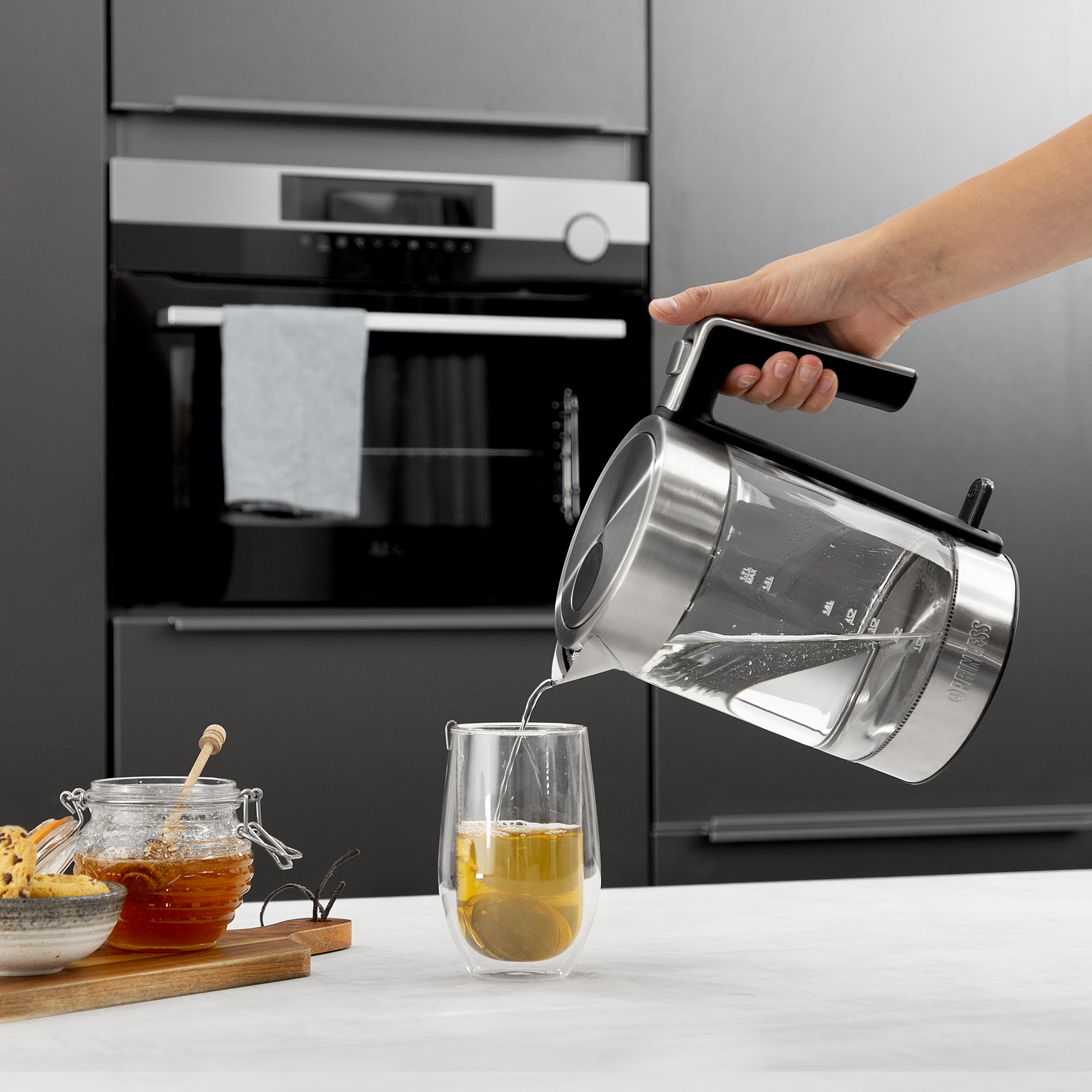 Hand gießt Wasser aus einem gläsernen Wasserkocher in ein Glas mit Teebeutel. Küchenszene.