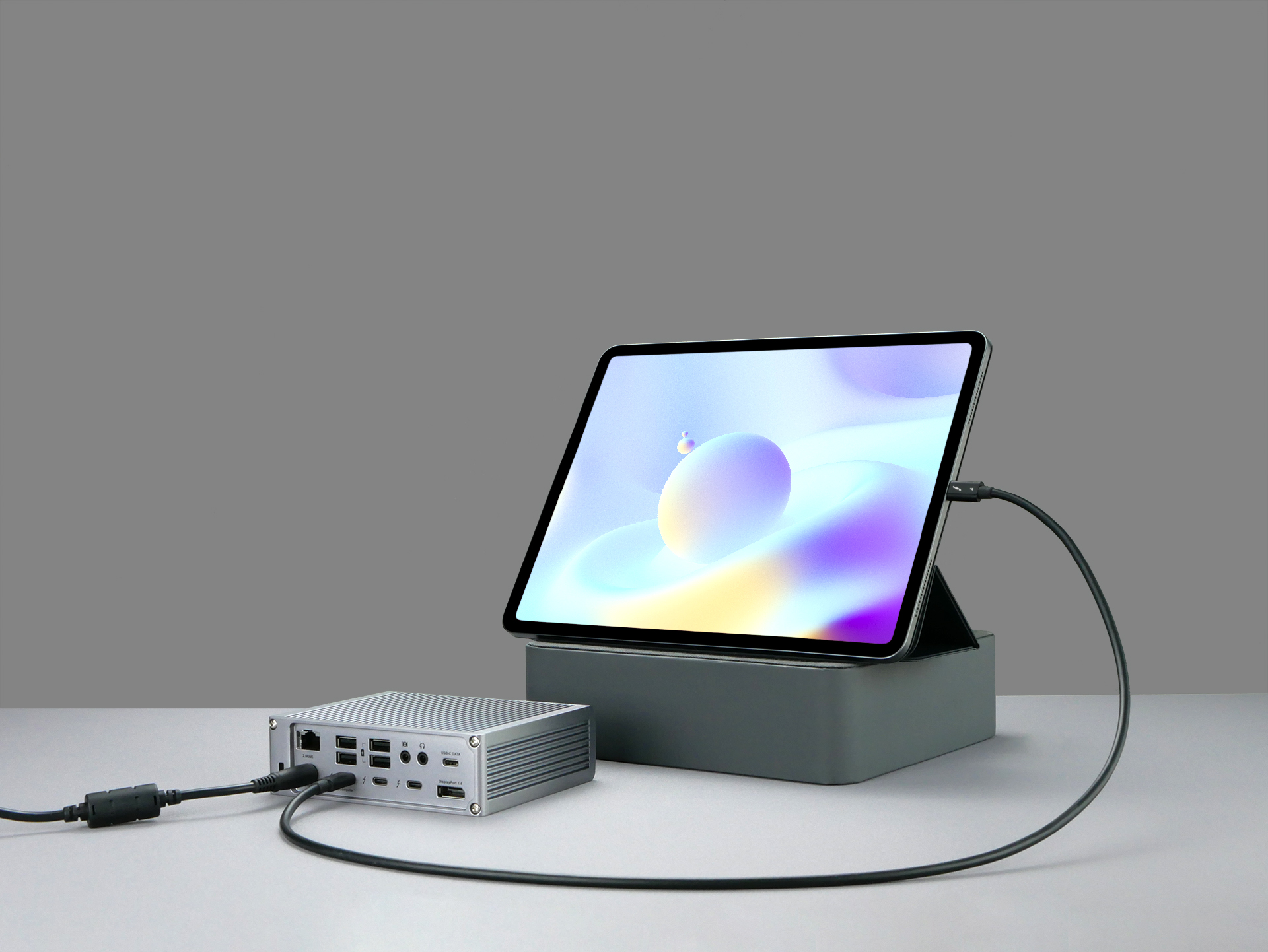Un tablet con un design astratto è collegato a un dispositivo argentato su una superficie grigia, cavo nero.
