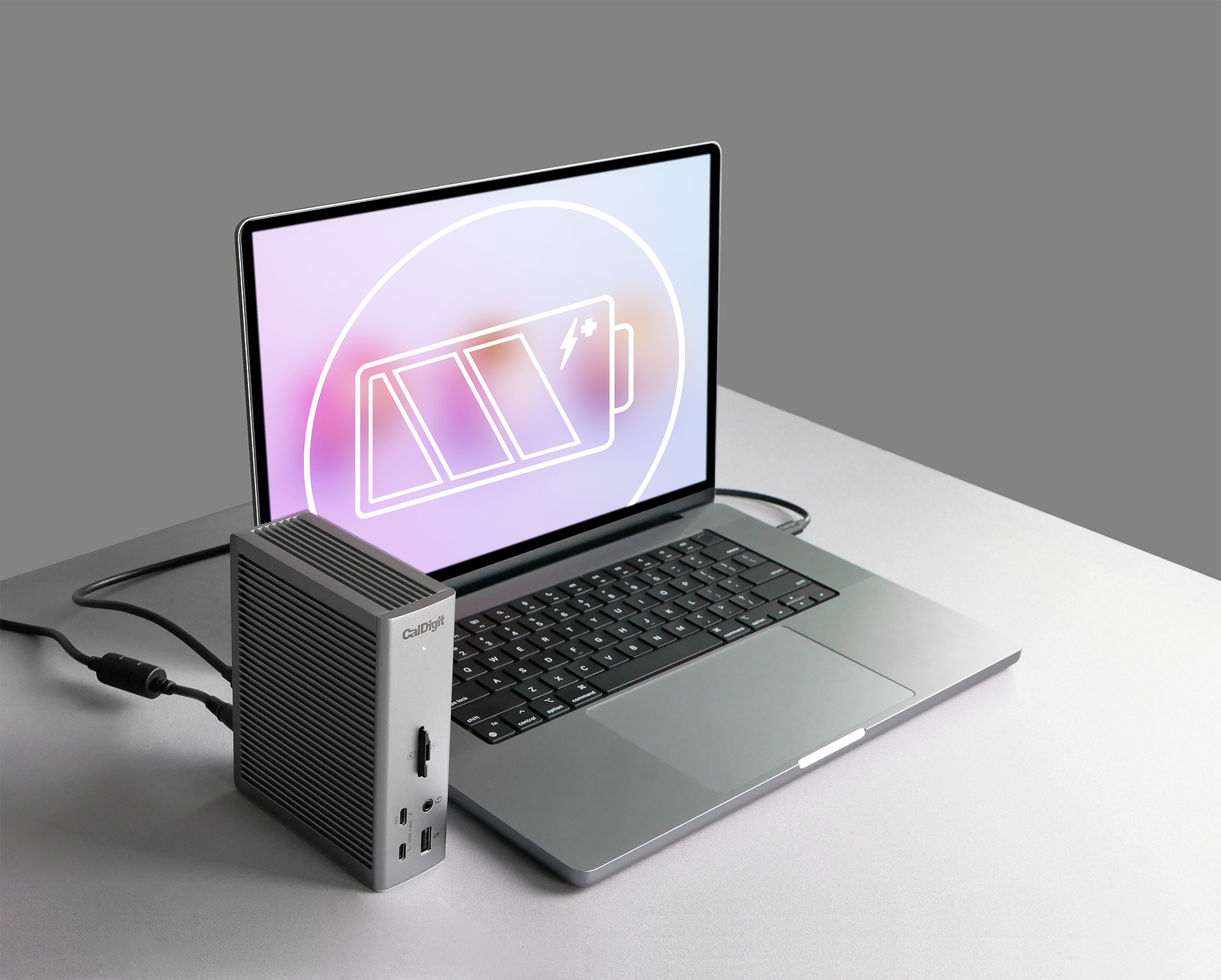 Laptop con simbolo di ricarica sullo schermo accanto a un dispositivo CalDigit grigio. Sfondo grigio.
