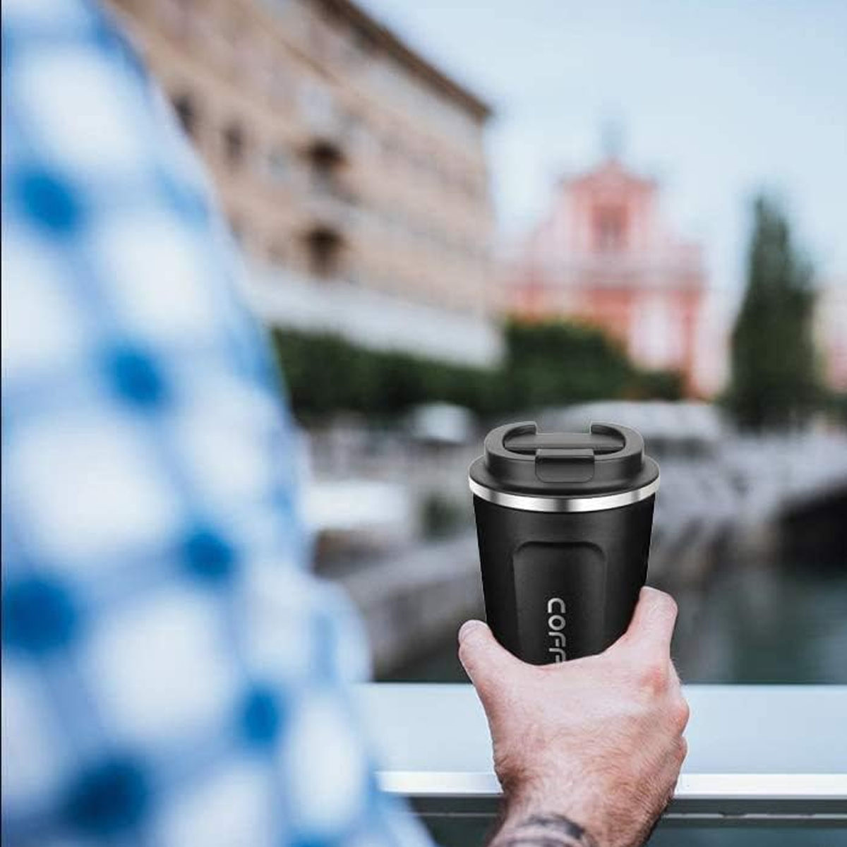 Person hält schwarzen Kaffeebecher mit Deckel, verschwommener Stadt im Hintergrund, Nahaufnahme.