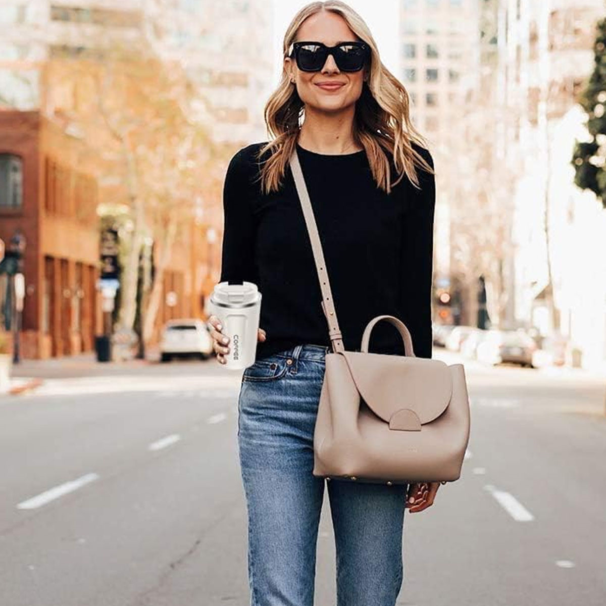 Eine Frau mit Sonnenbrille, die eine Kaffeetasse hält, steht auf einer Straße. Sie trägt Jeans und ein schwarzes Hemd.