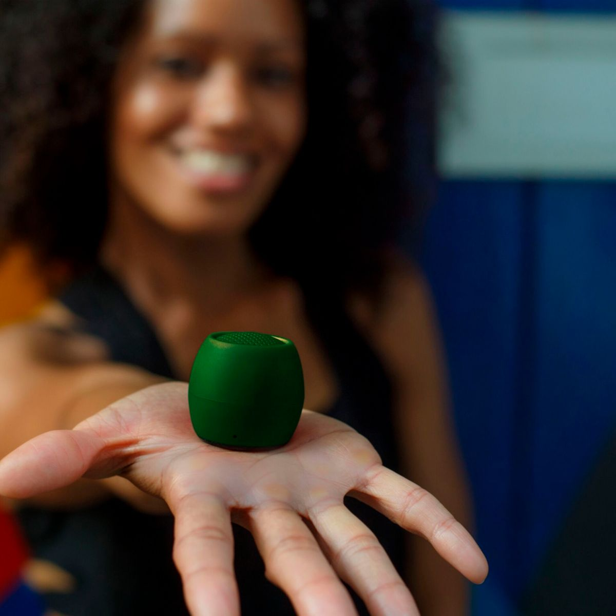 Kobieta trzymająca zielony głośnik. Uśmiechnięta, ciemne włosy. Głośnik jest zielony i okrągły.
