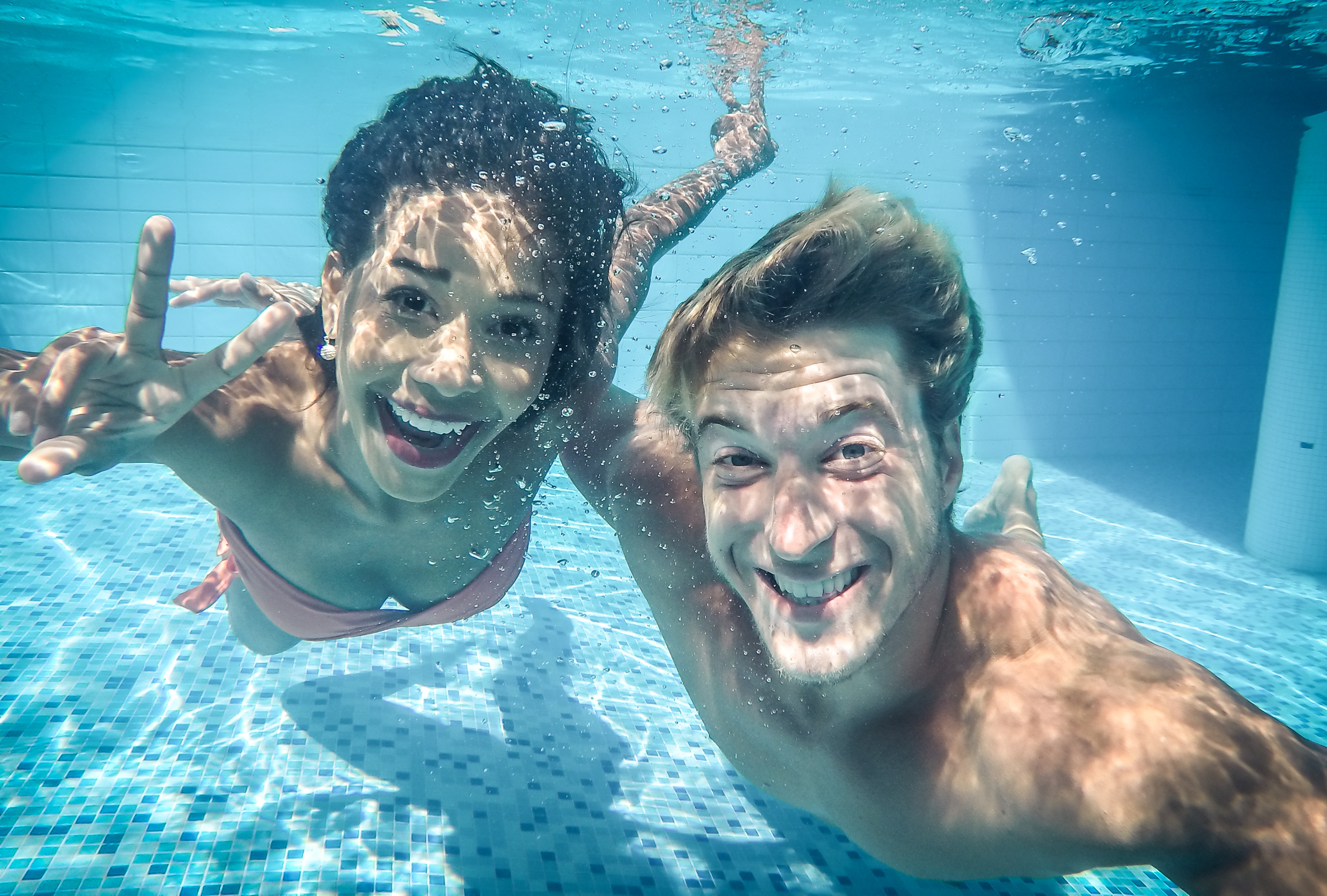 Zwei Menschen unter Wasser, lächelnd. Eine Frau macht ein Friedenszeichen. Blaues Wasser mit Sonnenreflexen.