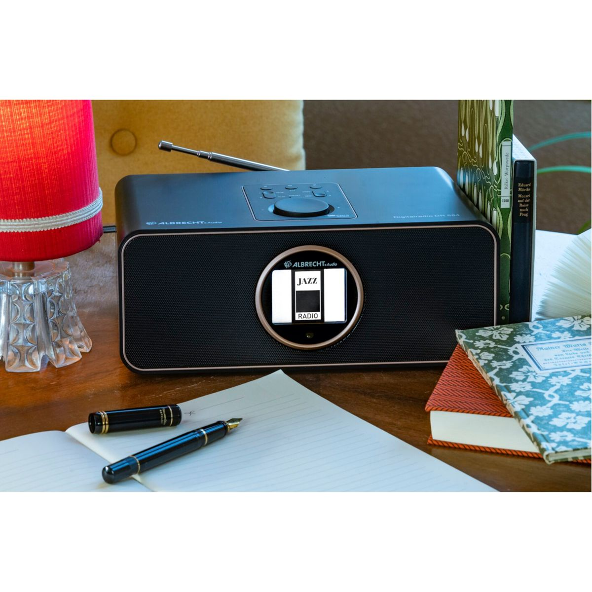 Une radio noire avec un affichage numérique, avec un logo Jazz Radio, sur un bureau avec des livres, des stylos et une lampe.