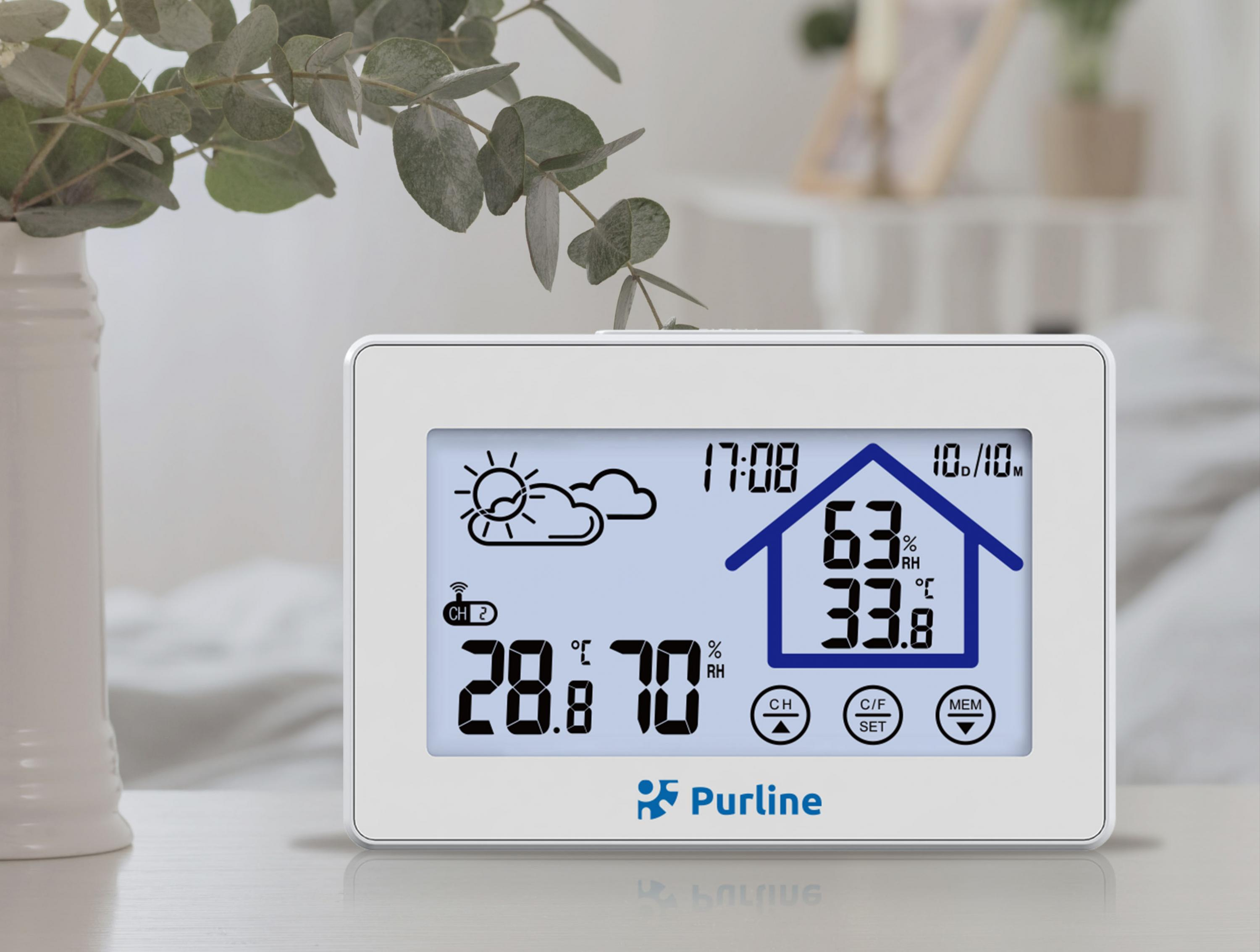Weiße Digitaluhr mit Zeit, Temperatur und Luftfeuchtigkeit. Vase und Pflanze im Hintergrund.